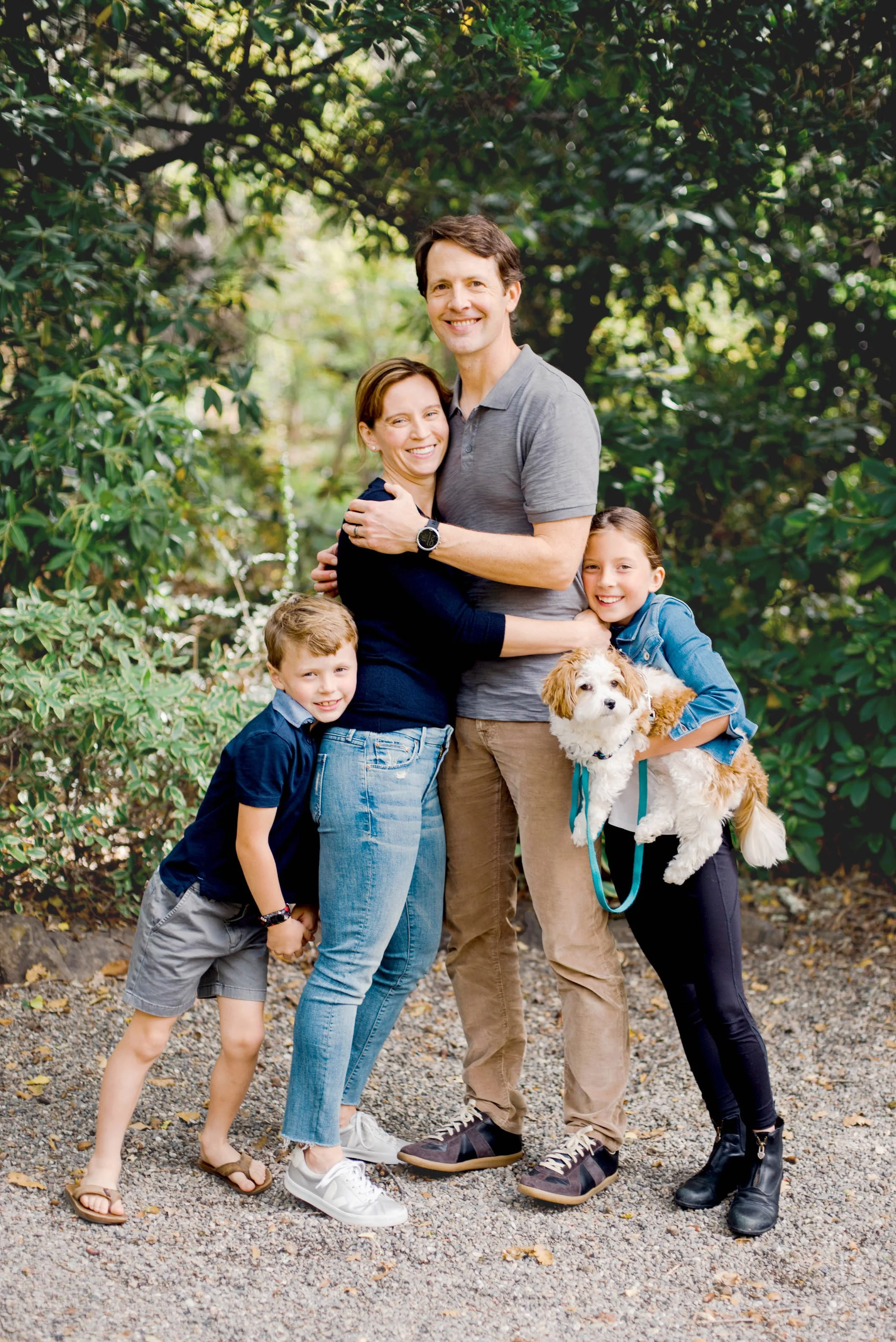 A family of five, including two young boys, a woman, a man, and a girl, along with a small dog, standing outdoors on a gravel path surrounded by green trees and foliage, smiling and embracing each other.