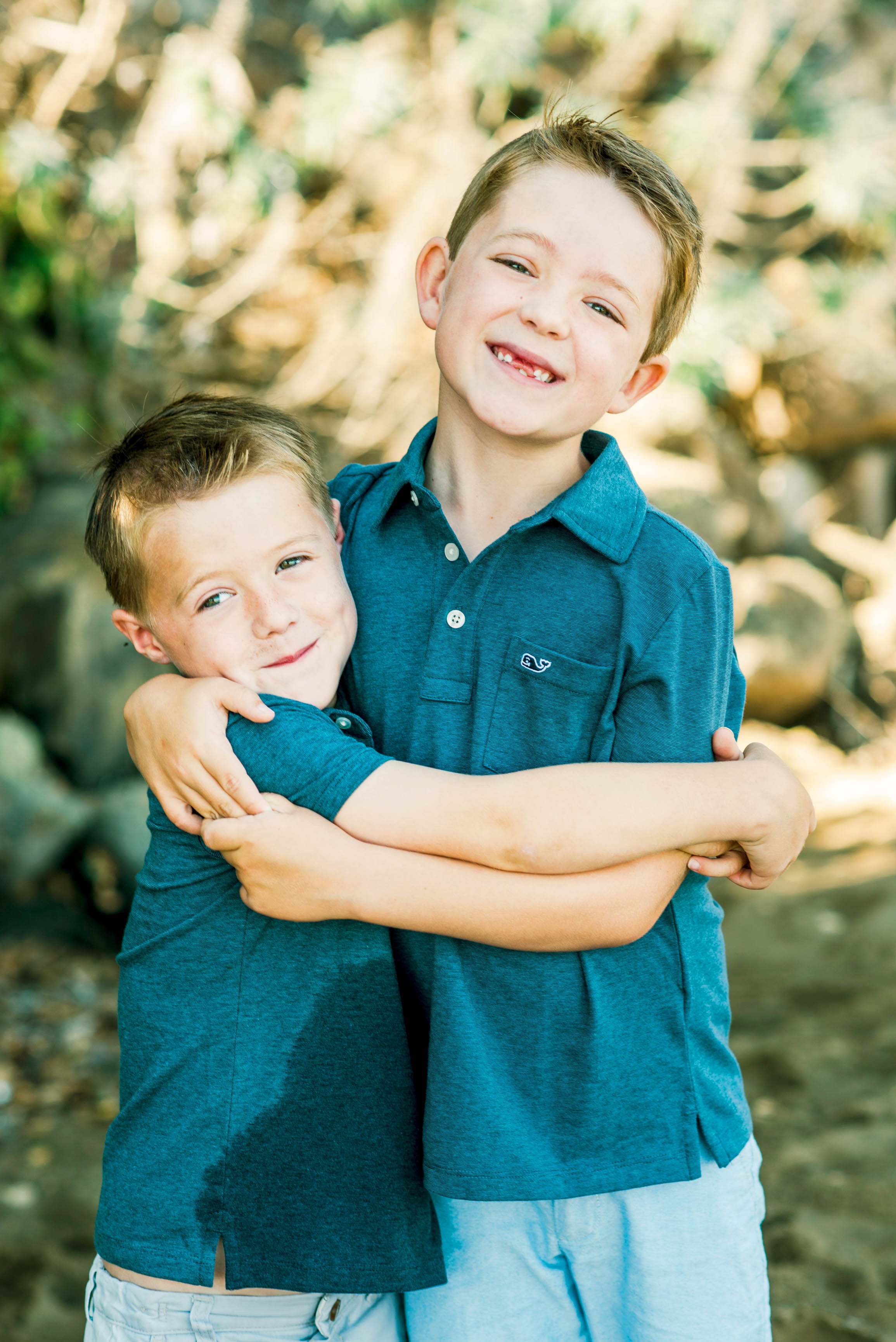 Two young boys wearing blue shirts, smiling and hugging outdoors.