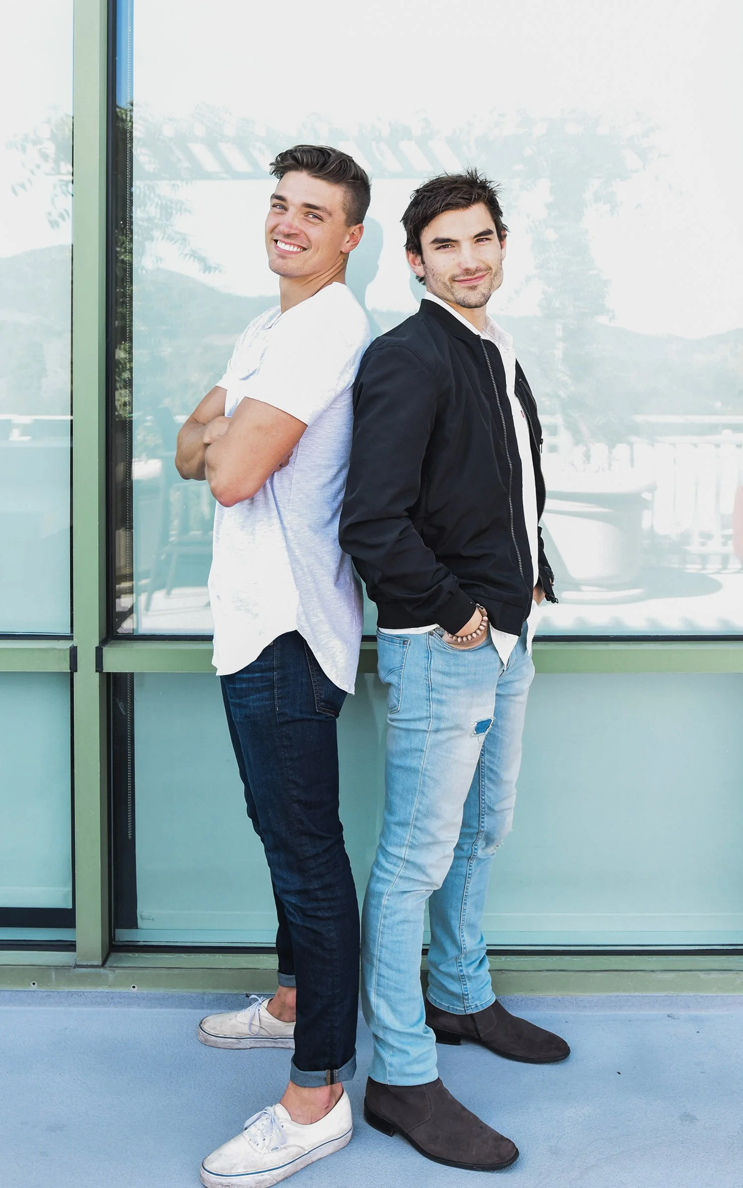 Two smiling men standing back-to-back in casual attire outside a glass building.