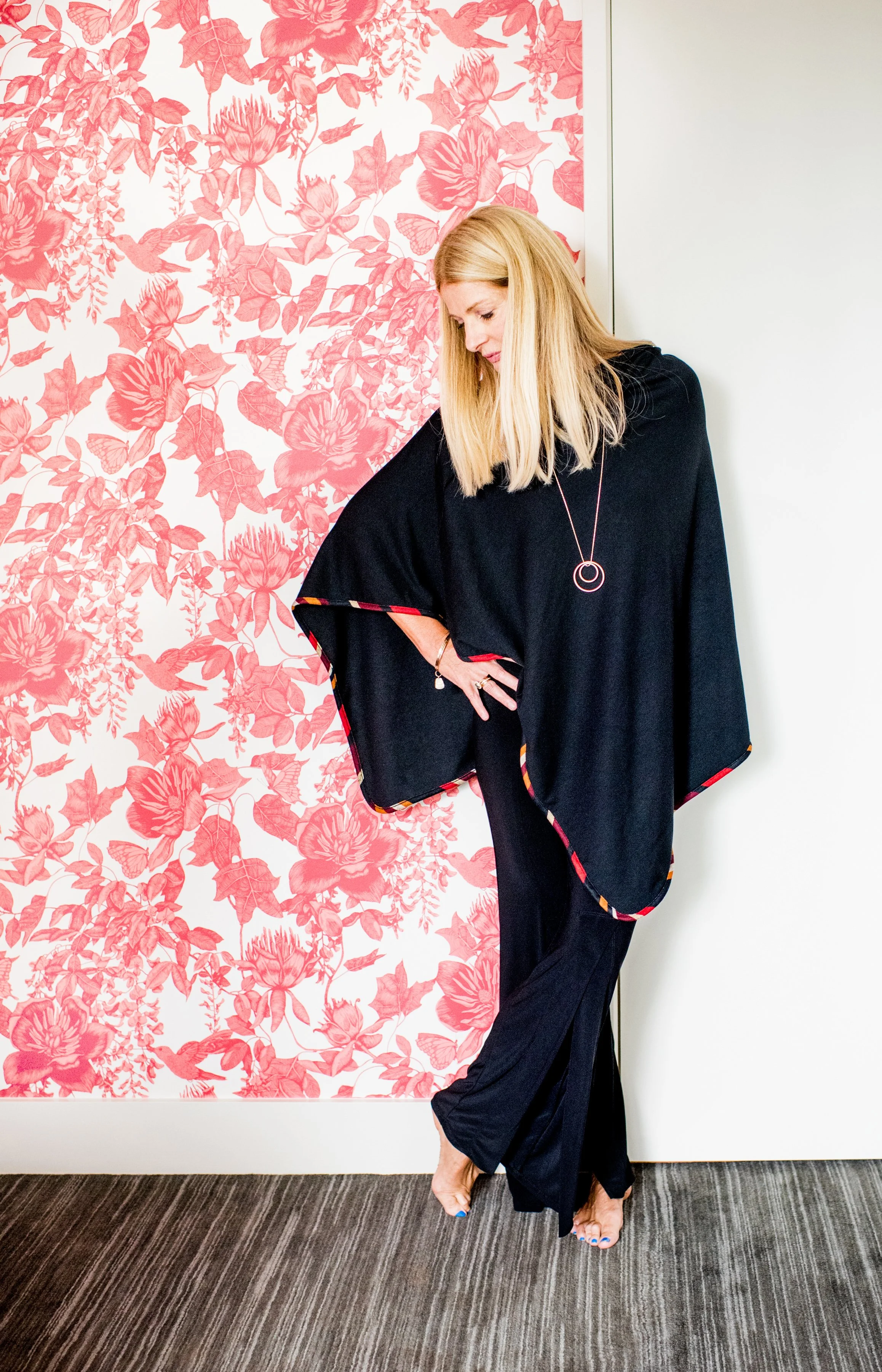 A woman with long blonde hair wearing a black dress and jewelry, standing on a striped carpet near a pink floral wallpaper and white wall.