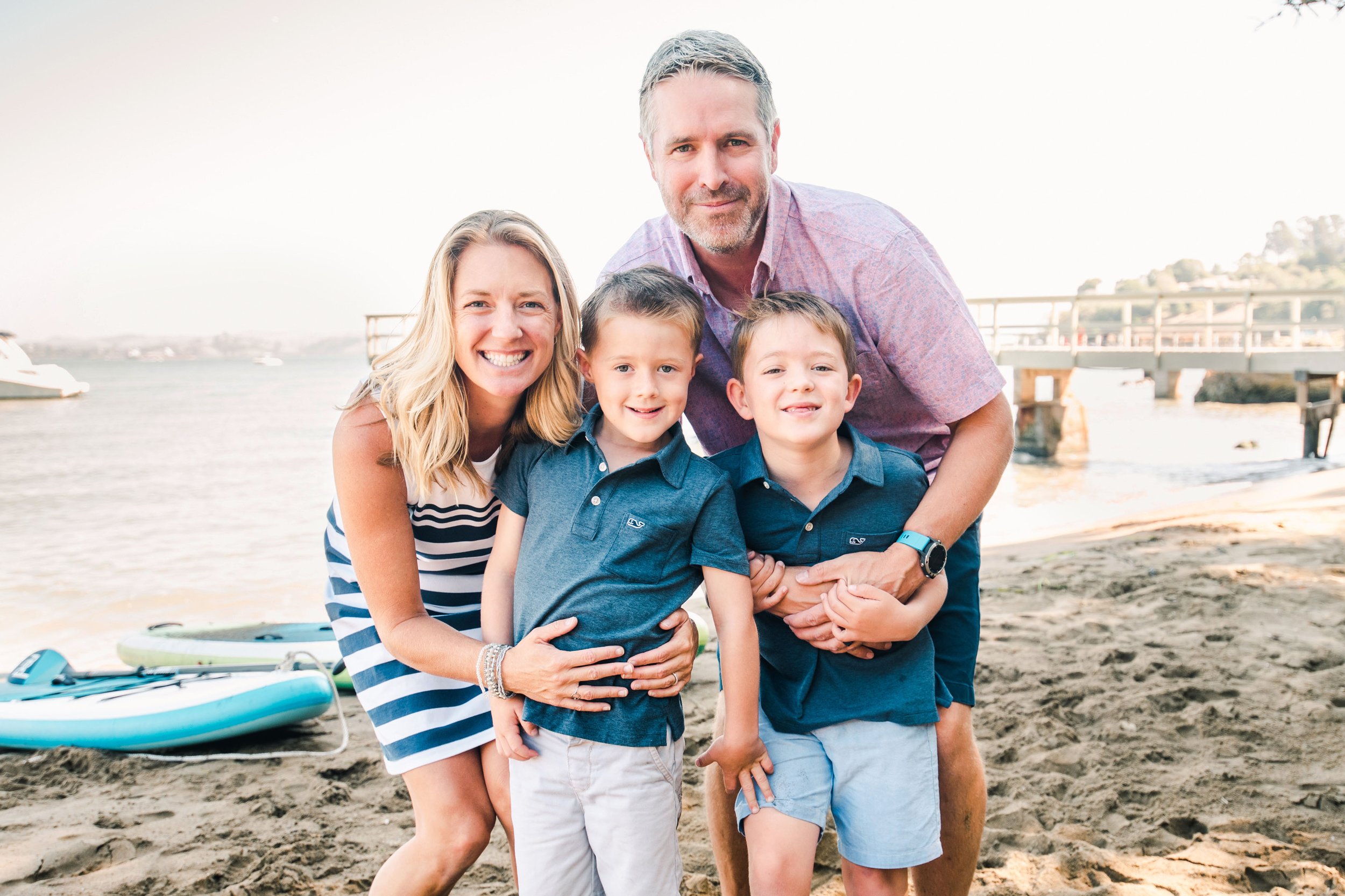 Family photo session in Tiburon, Ca, a family of four smiling on a sandy beach with water and paddleboards in the background.