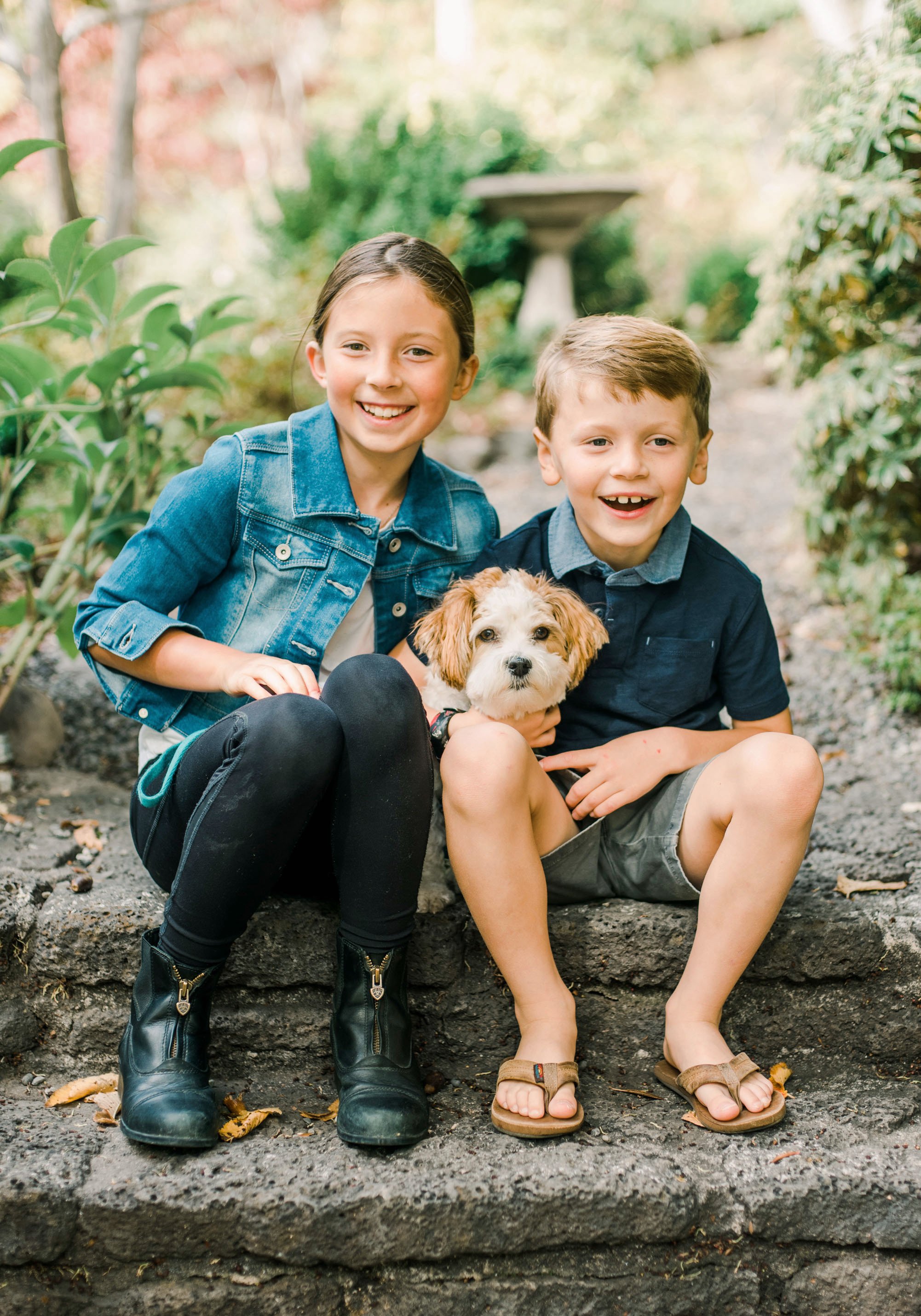 Two children, a girl and a boy, sitting on a stone ledge outdoors with a small, fluffy puppy. The girl is wearing a denim jacket, black pants, and black boots, smiling at the camera. The boy is in a dark shirt with a gray collar, beige shorts, and sandals, also smiling with the puppy on his lap. The scene is surrounded by greenery, with fall-colored trees in the background.