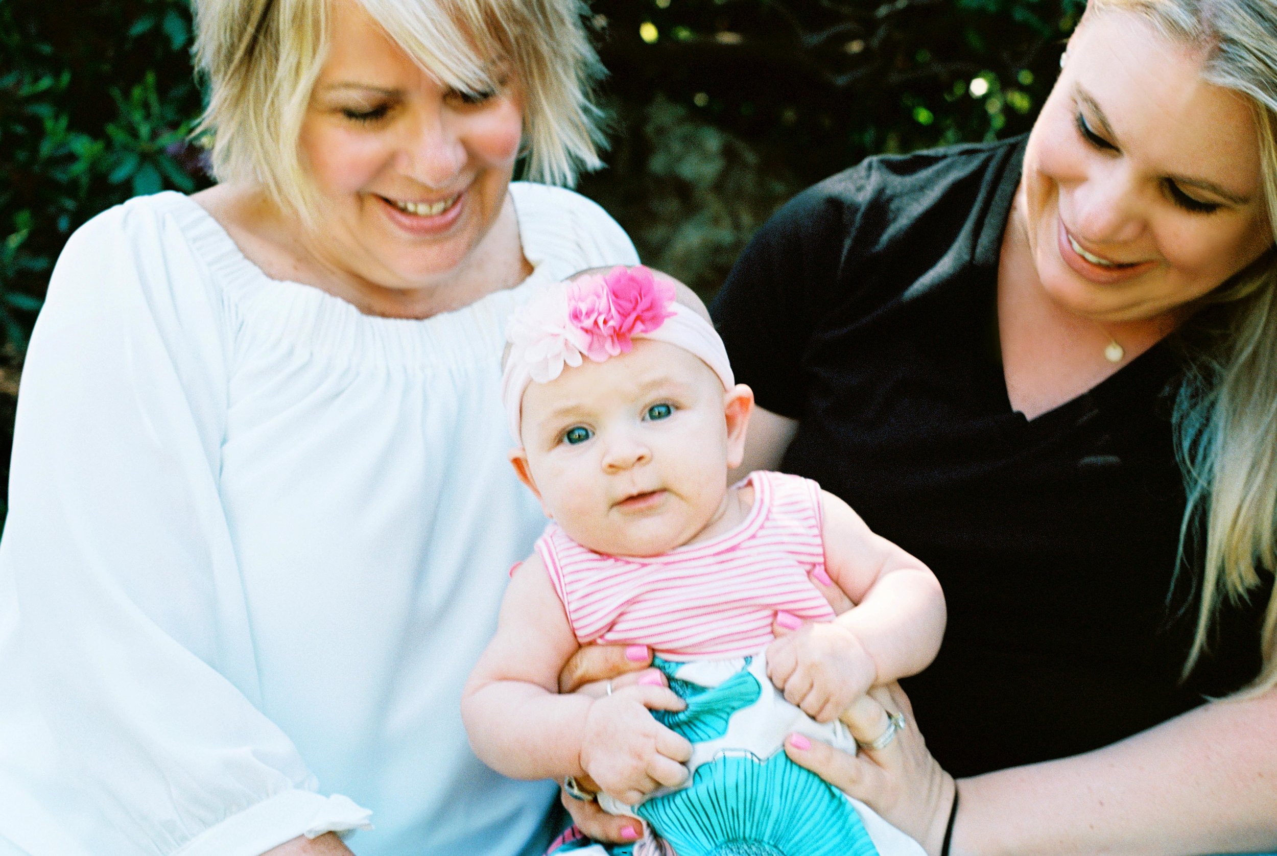 Three generations, two moms smiling at a baby girl wearing a flower headband and striped dress.