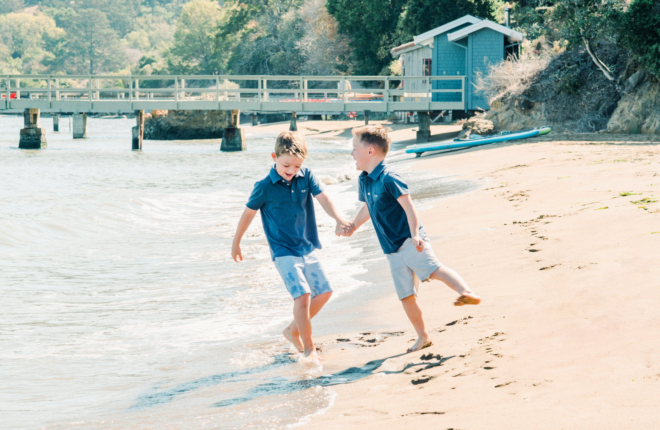 Two young boys play on a sandy beach near a pier and a small blue building, with one boy kicking water as they hold hands.