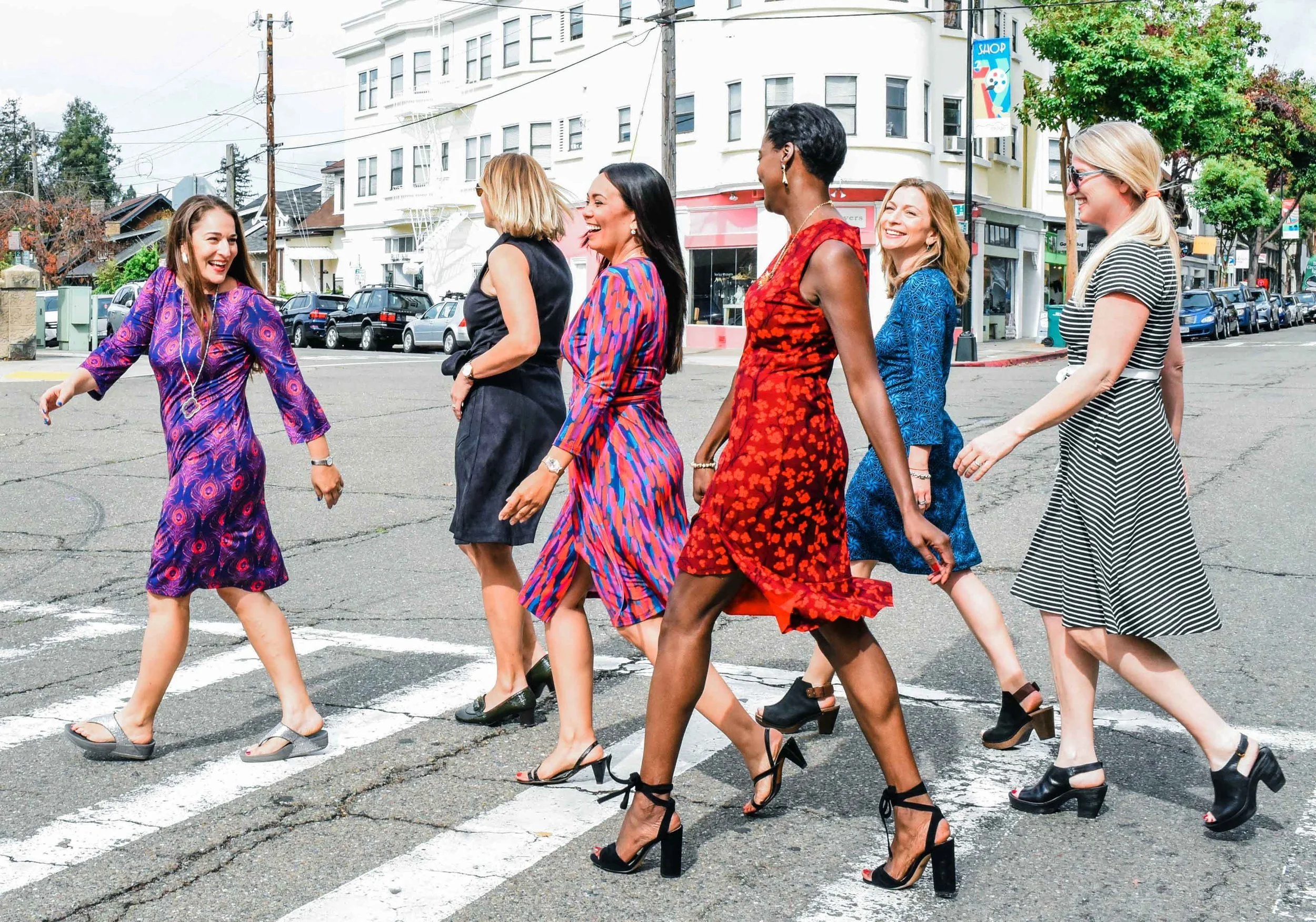 A group of women walking across a crosswalk in an urban area, smiling and enjoying each other's company. They are dressed in colorful dresses and high heels, and the background features buildings, parked cars, and trees.