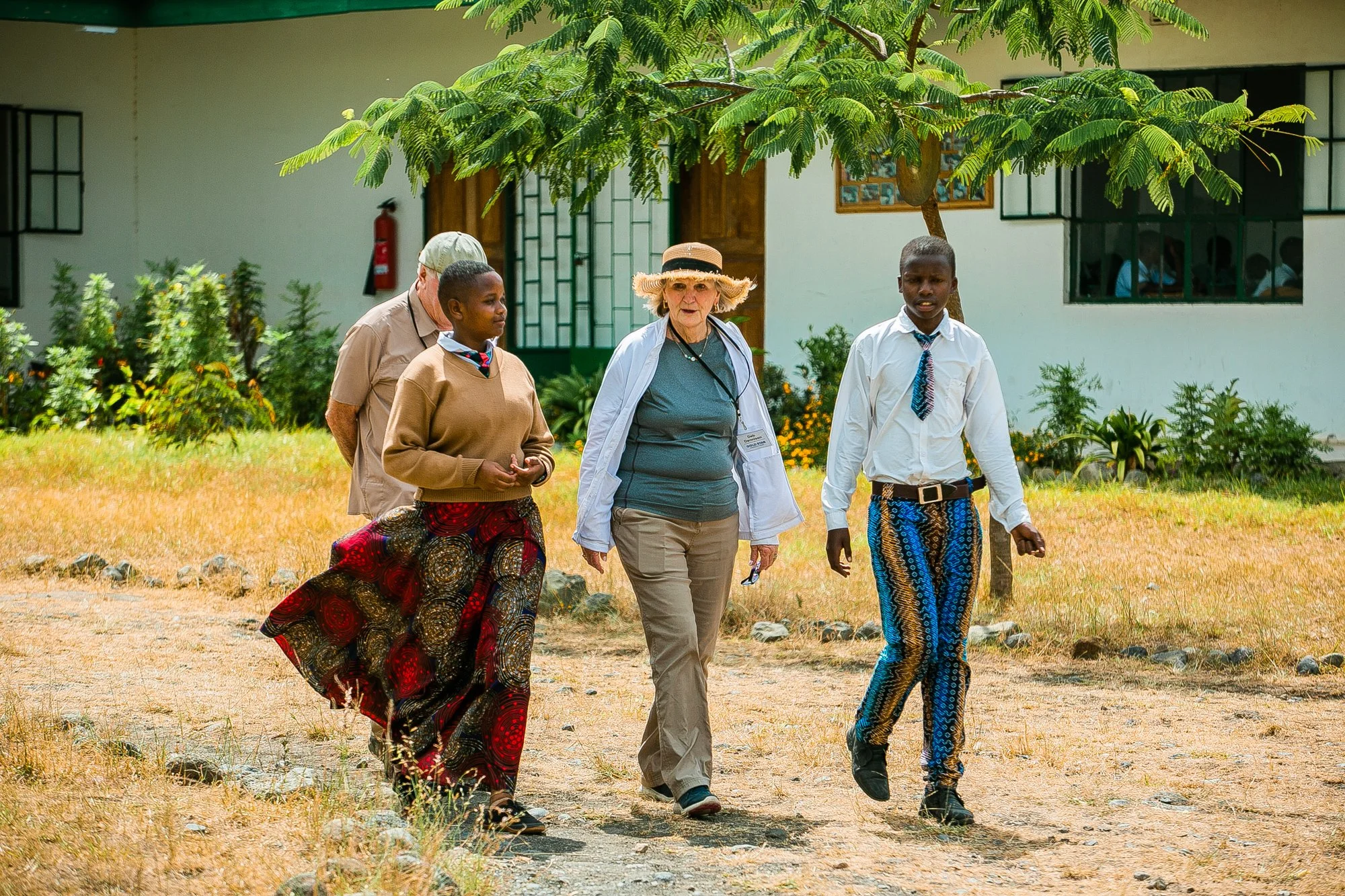 A diverse group of four people walking outdoors on a sunny day, with a white building, windows, and green foliage in the background. One woman is wearing a wide-brimmed hat and white jacket, two young men are dressed in traditional African patterned pants and white shirts, and an older man is behind them.