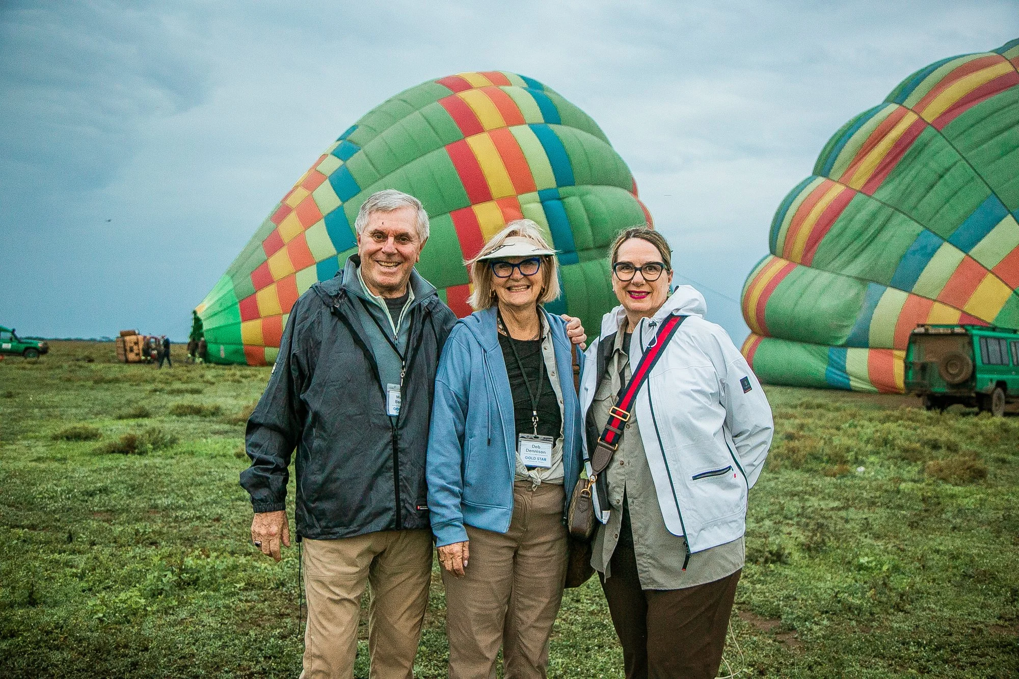 Three people standing outdoors in front of colorful hot air balloons on a grassy field, with a cloudy sky above.