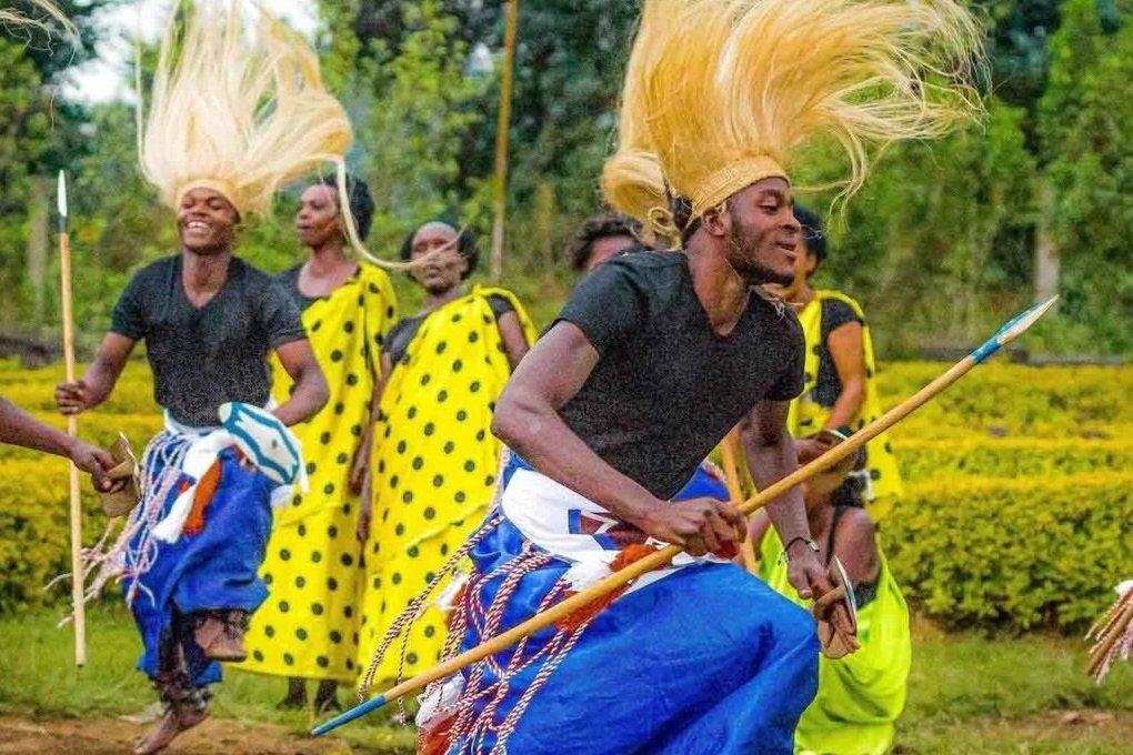 Group of people in Rwanda, in colorful traditional attire dancing outdoors with greenery in the background, some holding spears and wearing tall, golden hair accessories.