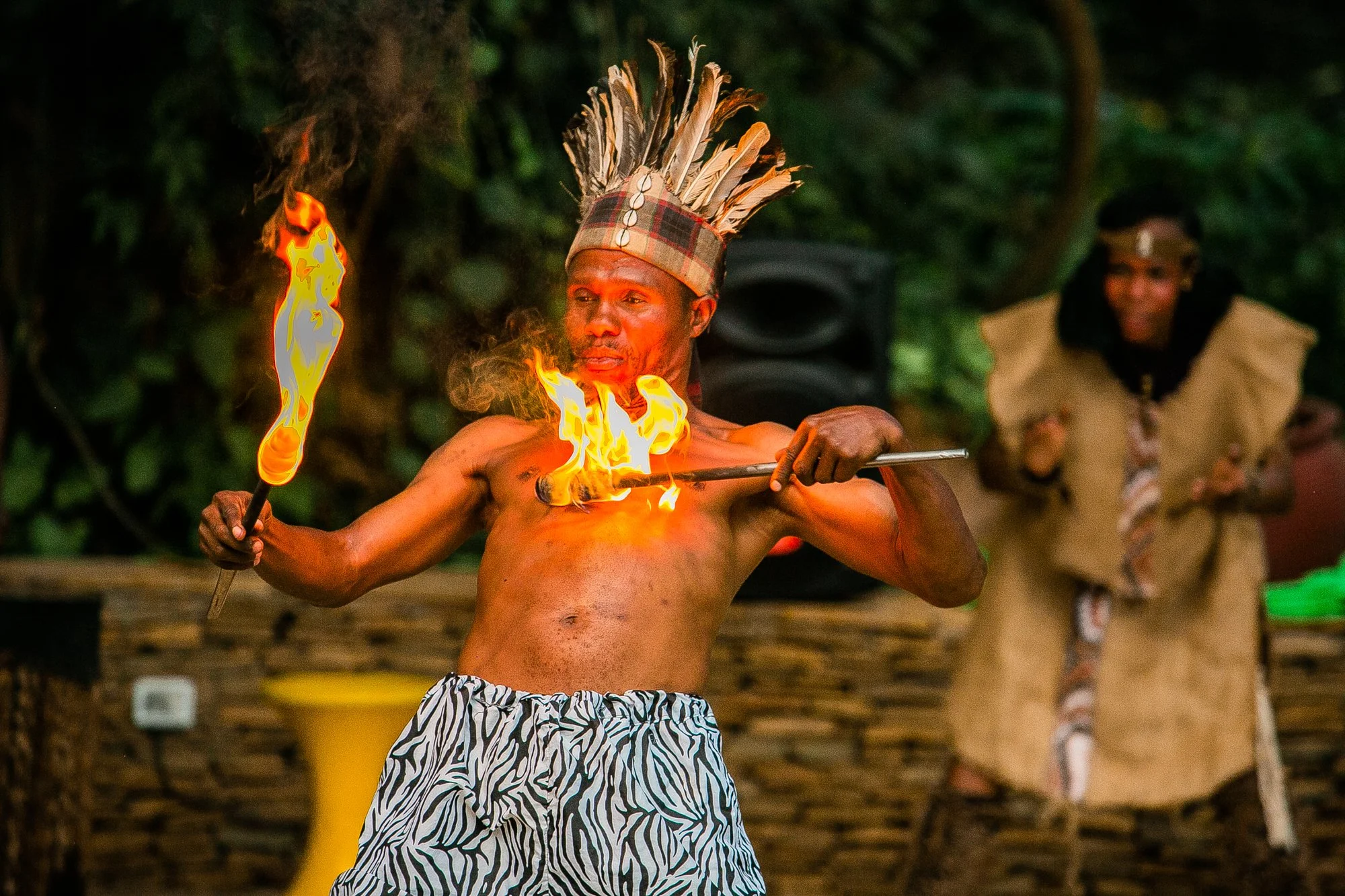 A man performing a fire dance in traditional attire, wearing a headdress made of feathers and zebra-patterned shorts, holding a flaming stick at night in a forest setting. Another person is blurred in the background.