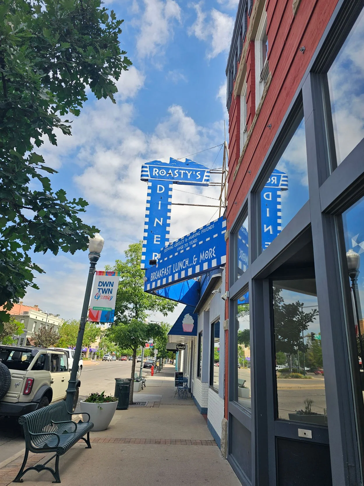 Street view showing a blue sign for Roasty's Diner advertising breakfast, lunch, and more, on a sunny day with a few clouds, trees, parked cars, and a sidewalk with benches and planters.