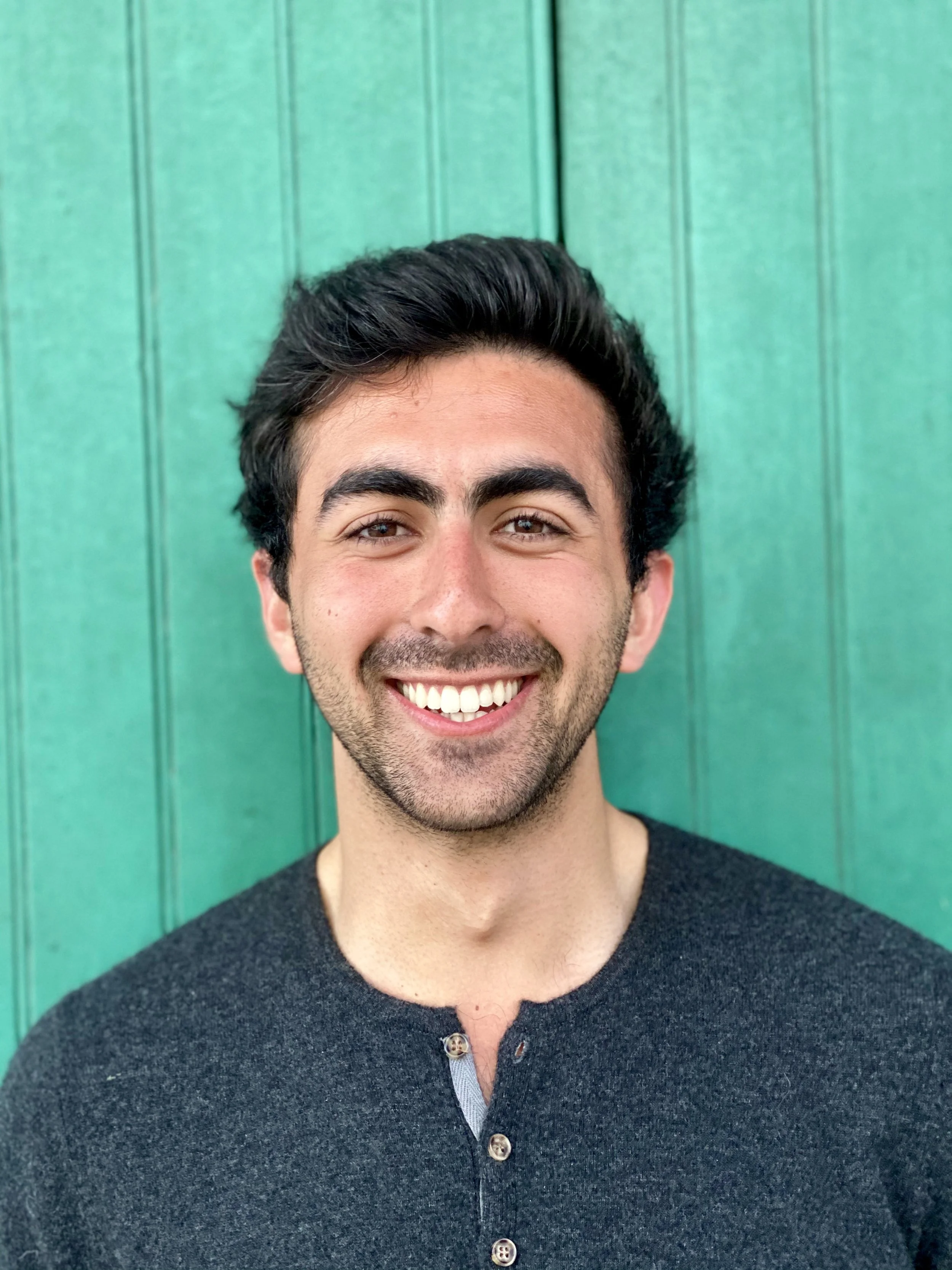 Young man with dark hair and a beard smiling, wearing a dark buttoned shirt, standing in front of a green wooden background.