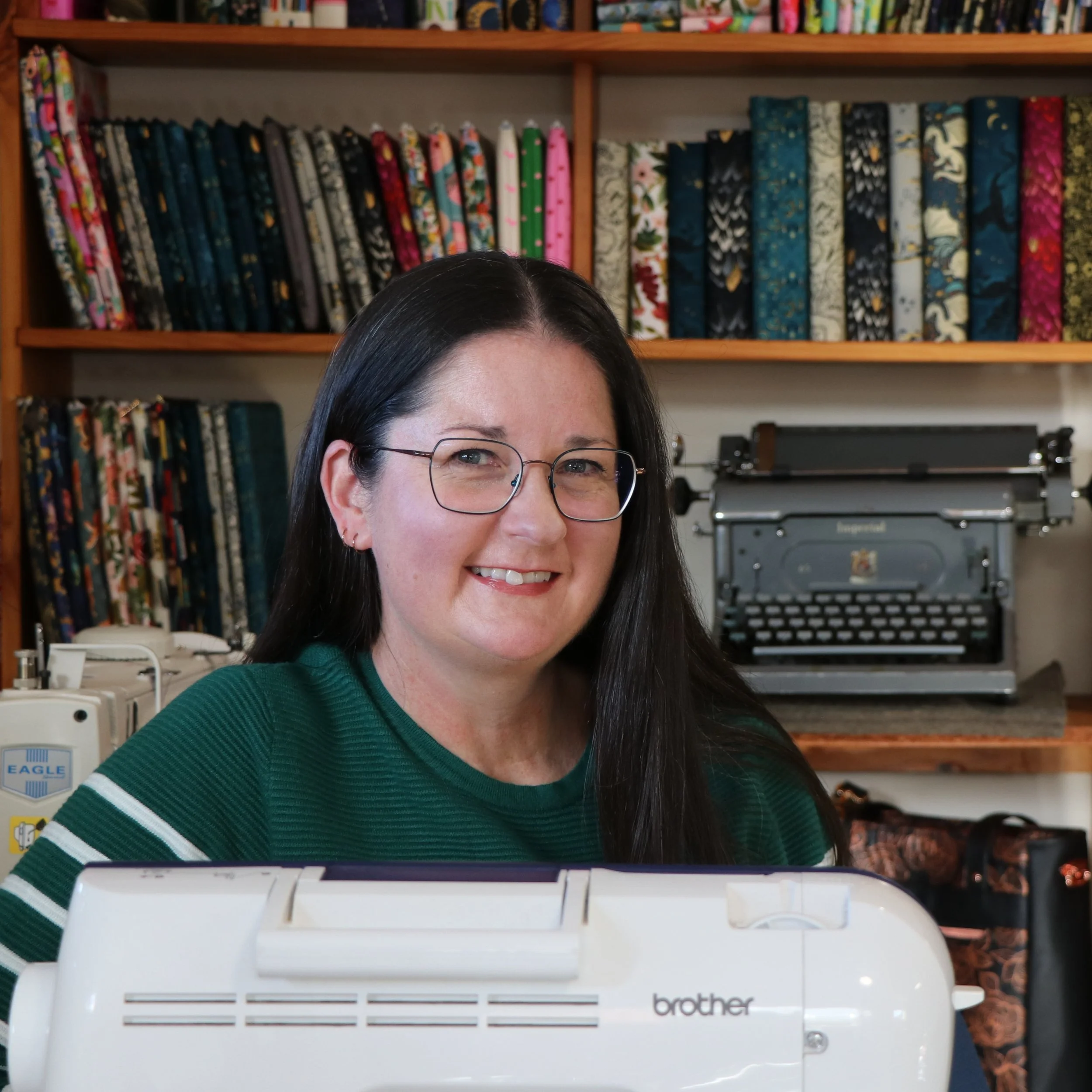 Karen, owner of Kaybee Creations, sitting behind a sewing machine in her home studio with handmade book sleeves, fabrics and a vintage typewriter displayed behind her.