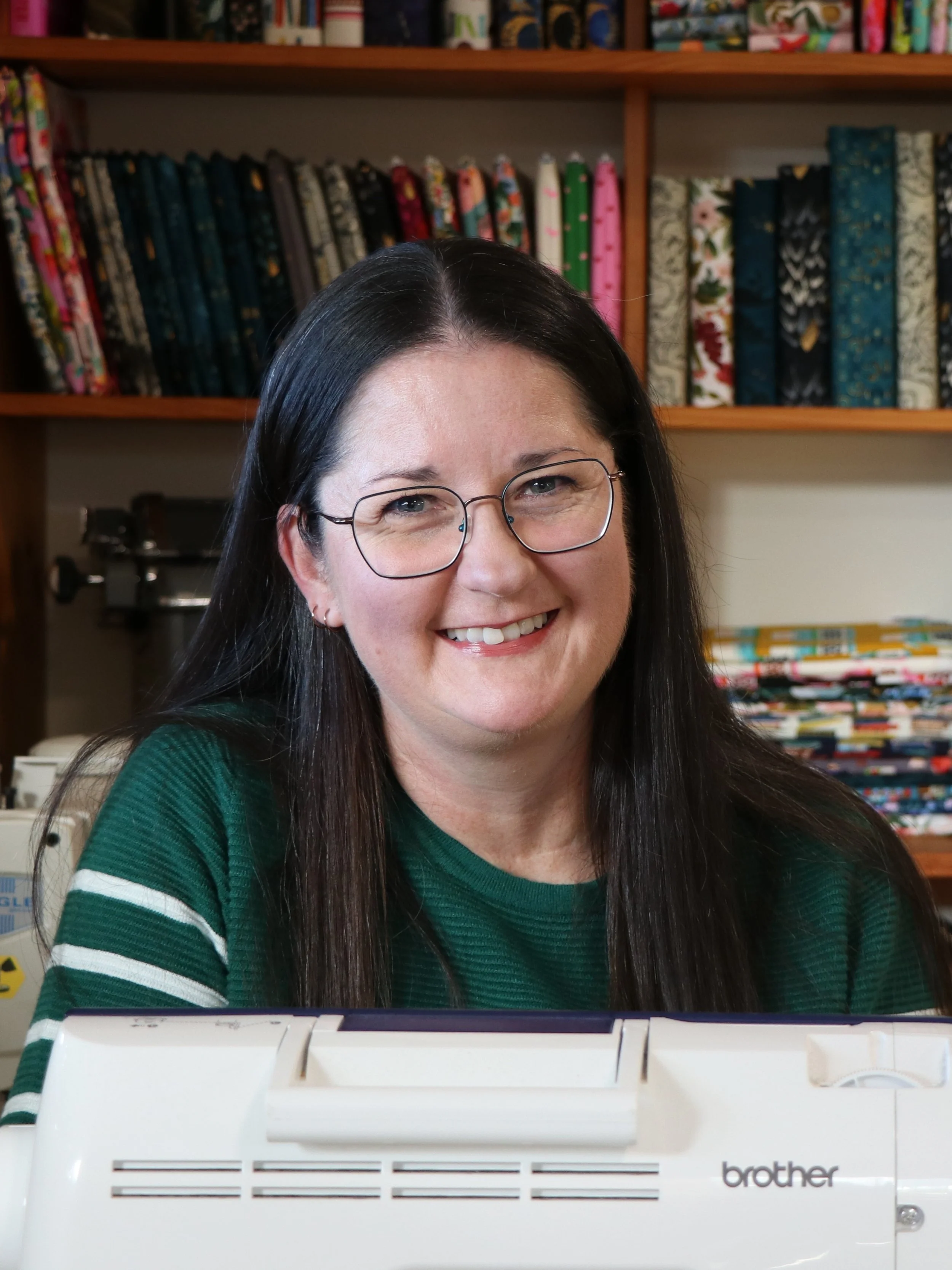 Karen, maker behind Kaybee Creations, smiling behind a sewing machine in her sewing studio with shelves of handmade book sleeves and colourful fabrics in the background.