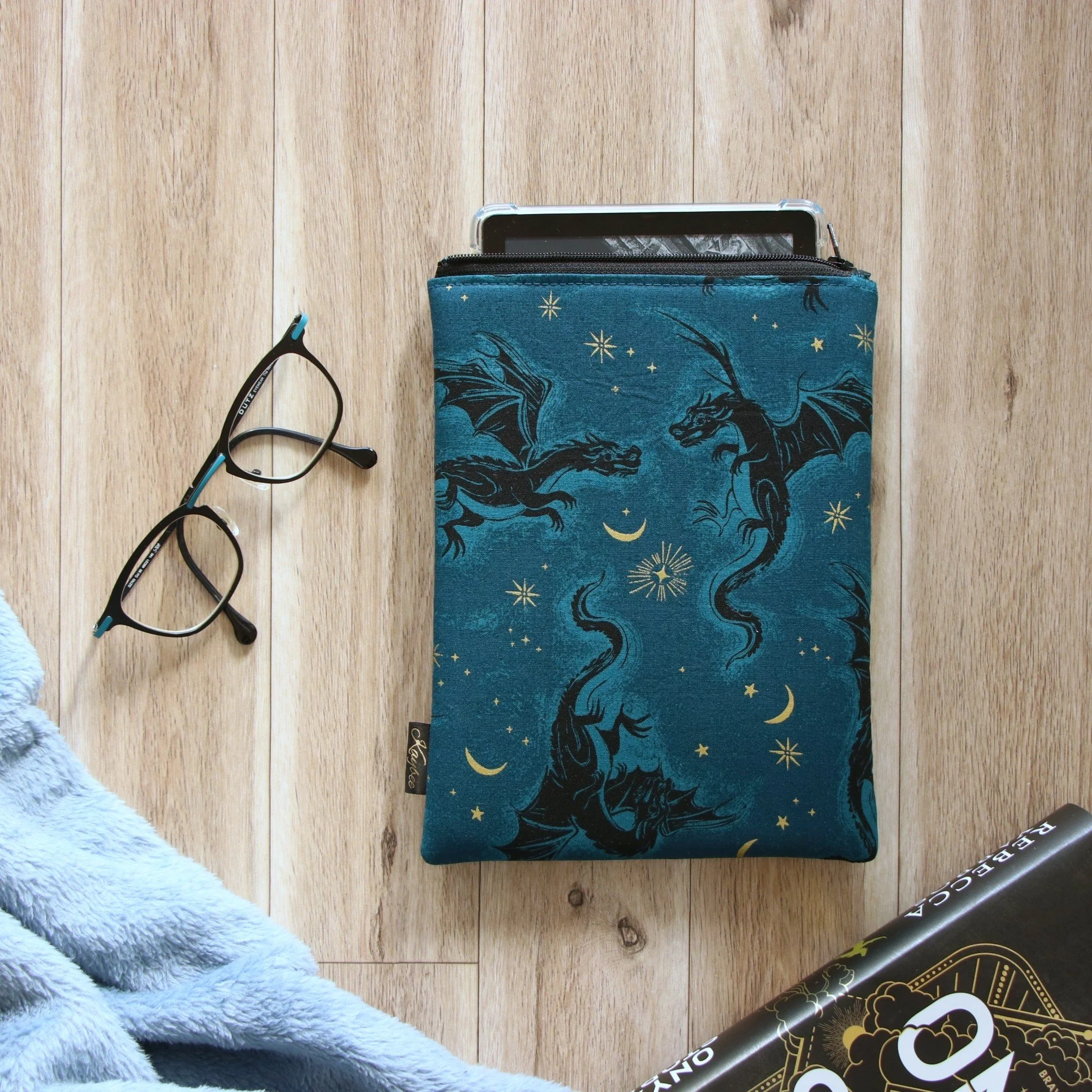 Handmade First Edition Kindle Sleeve with dragon-inspired fabric beside book and reading accessories.