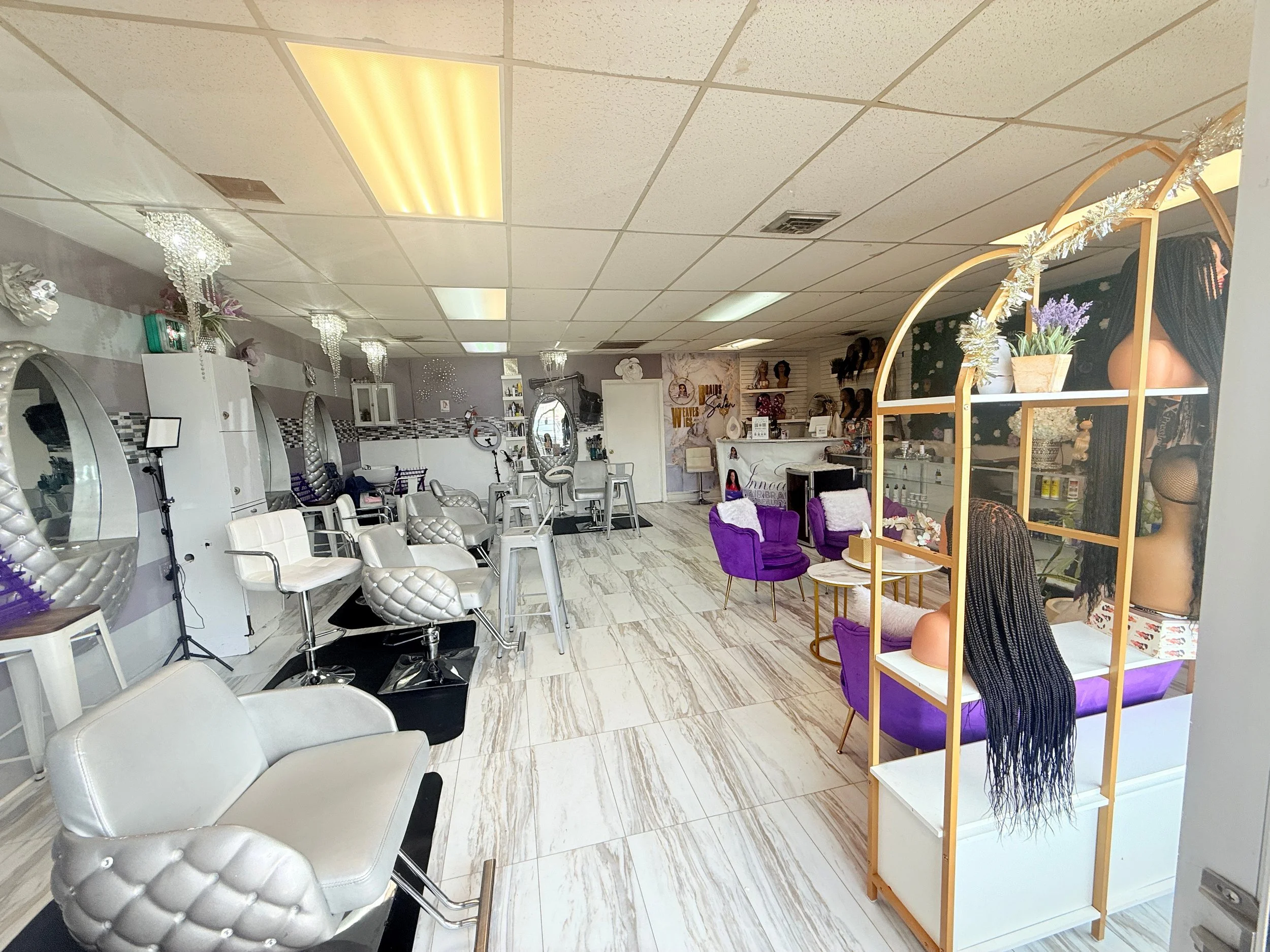 Interior of a hair salon with styling chairs, mirrors, and hair products, decorated with purple chairs, white and gold accents, and mannequins with wigs.