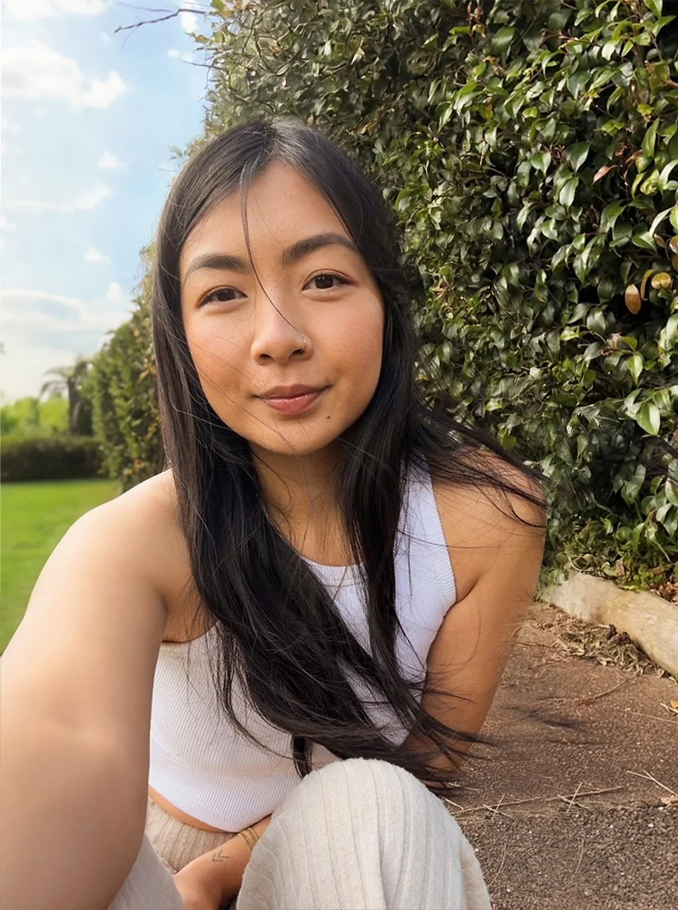 Young woman with long black hair taking a selfie outdoors beside a green hedge on a sunny day.