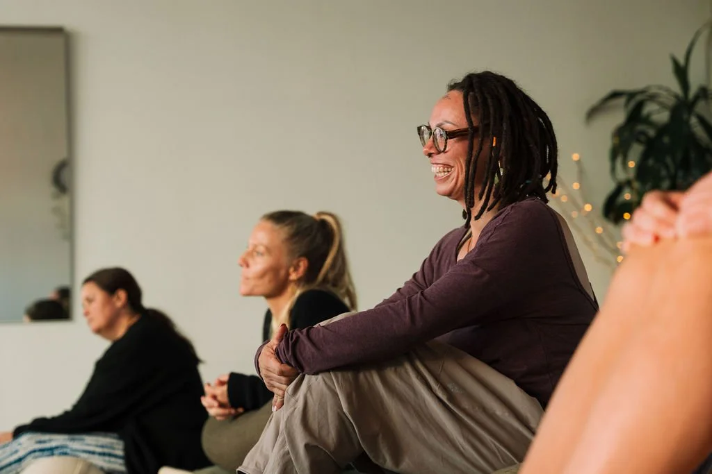 Group of women sitting cross-legged during a meditation or mindfulness session in a room with a white wall and a potted plant.