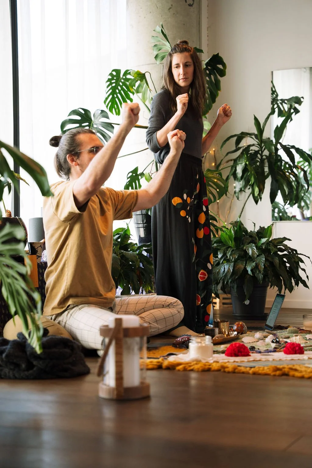 A man and a woman in a cozy indoor space with large green plants, one sitting on a cushion and the other standing, showing gestures with their hands to explain tetany in the hands during Breathwork.