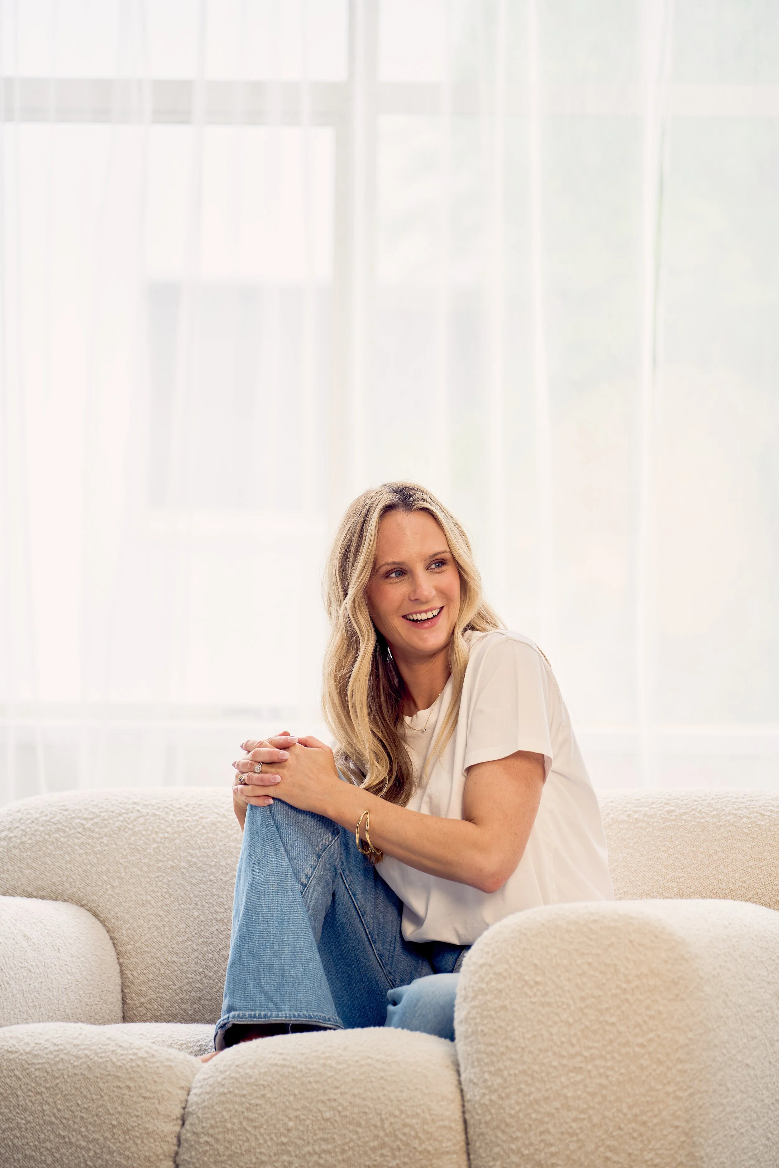 A woman sitting on a beige couch, smiling and looking to her right, with a bright window with curtains behind her.