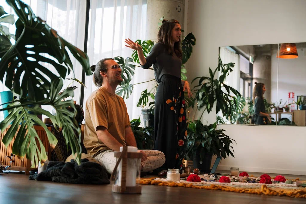 A woman standing and speaking with her hand raised, and a man sitting cross-legged on the floor smiling at a group who are listening to them, in a room with large plants, a mirror, and a colorful rug.