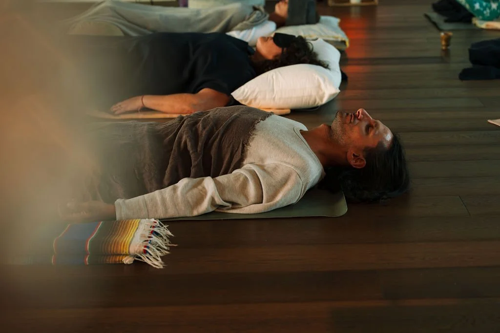 Two people are lying down on yoga mats, breathing slowly in a peaceful indoor space.