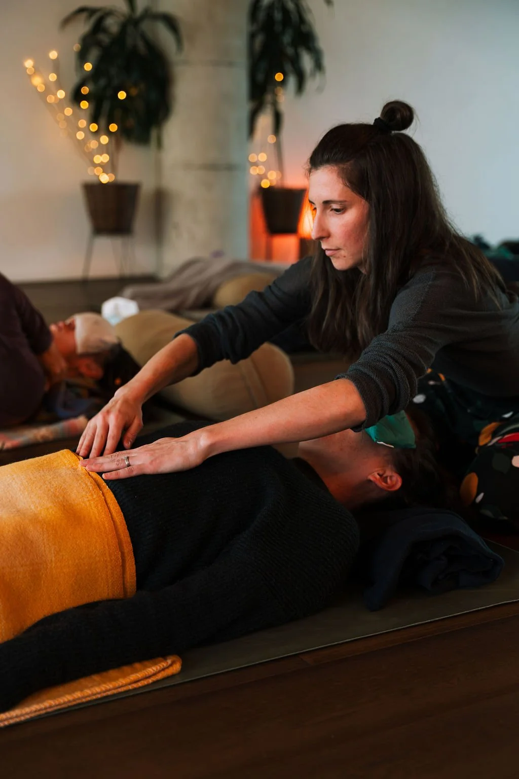 A female breathwork facilitator assiting a person lying on a mat in a cozy indoor setting with potted plants and warm lighting.