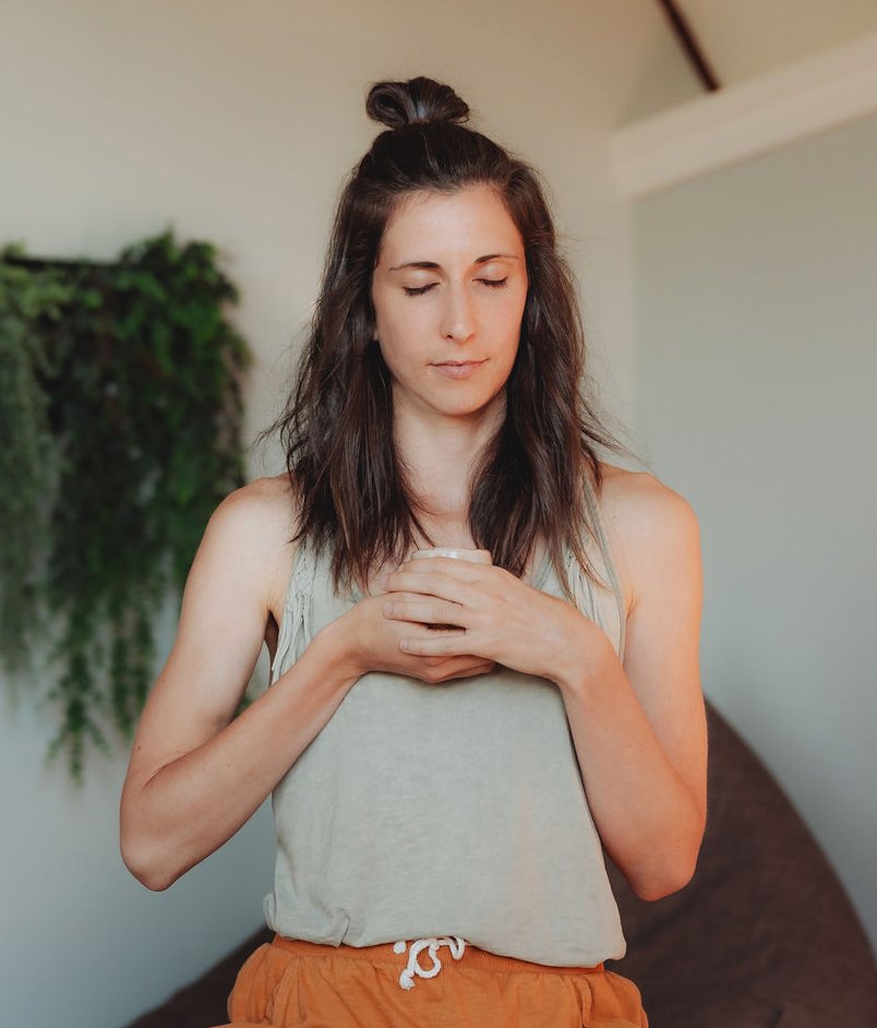 Woman with closed eyes, holding her hands over her chest, standing indoors.