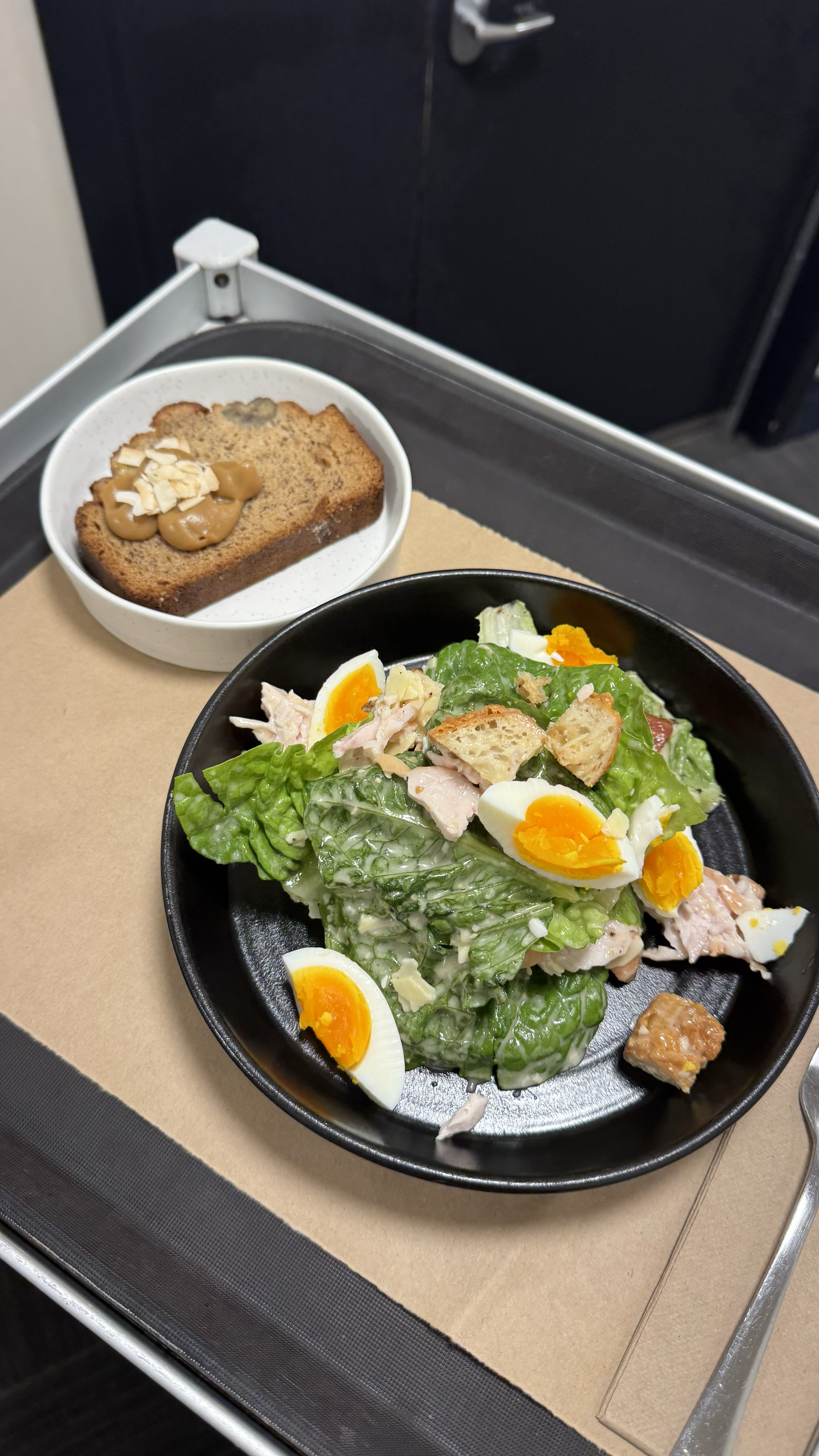 A tray with a black bowl of Caesar salad with romaine lettuce, hard-boiled eggs, croutons, and dressing, and a small bowl of banana bread topped with almonds in an airplane meal setting.