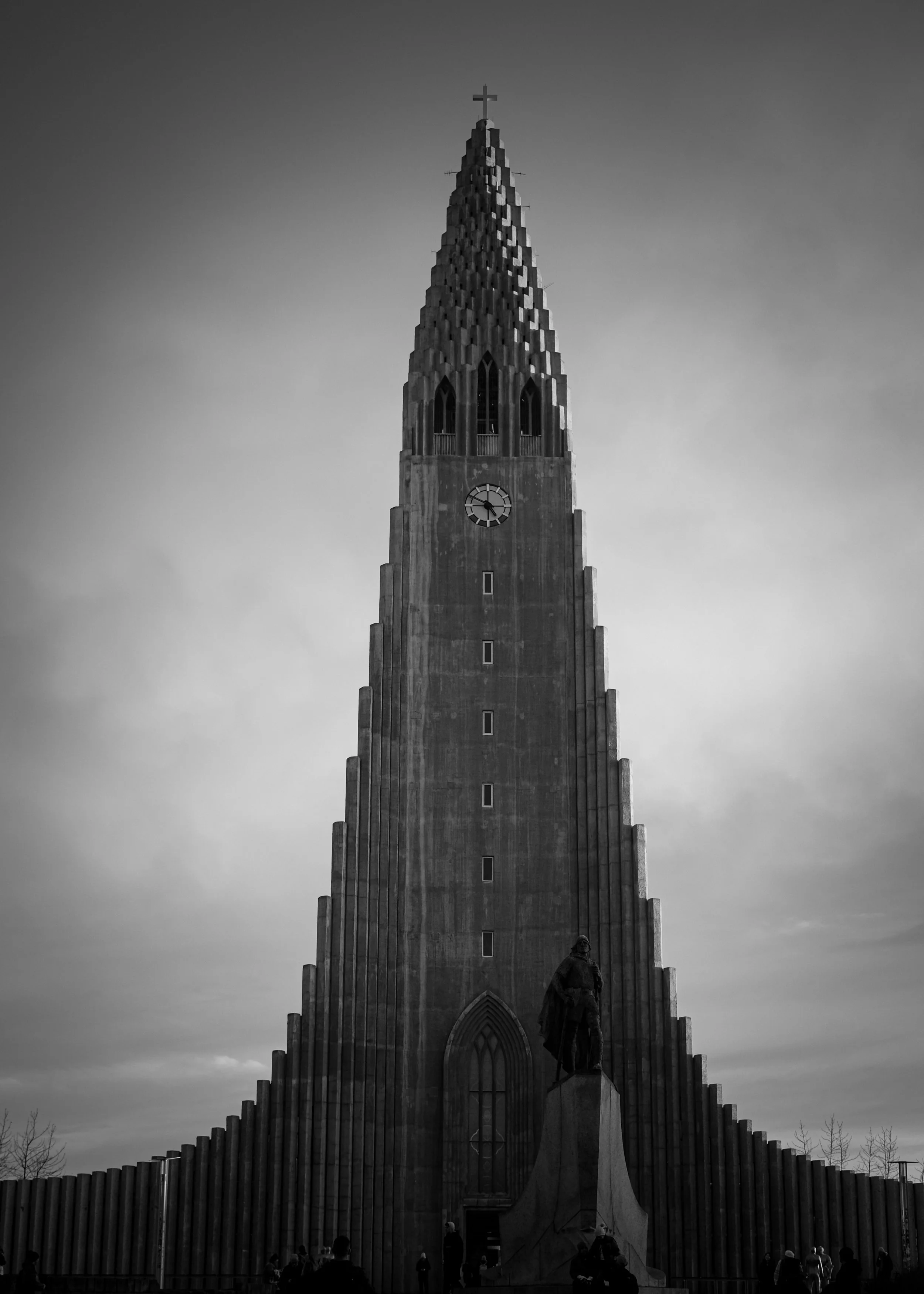 The Latter-day Saint temple, featuring a high, pointed spire topped with a cross, with a large clock and statues in front, surrounded by a low wall with several people in front.