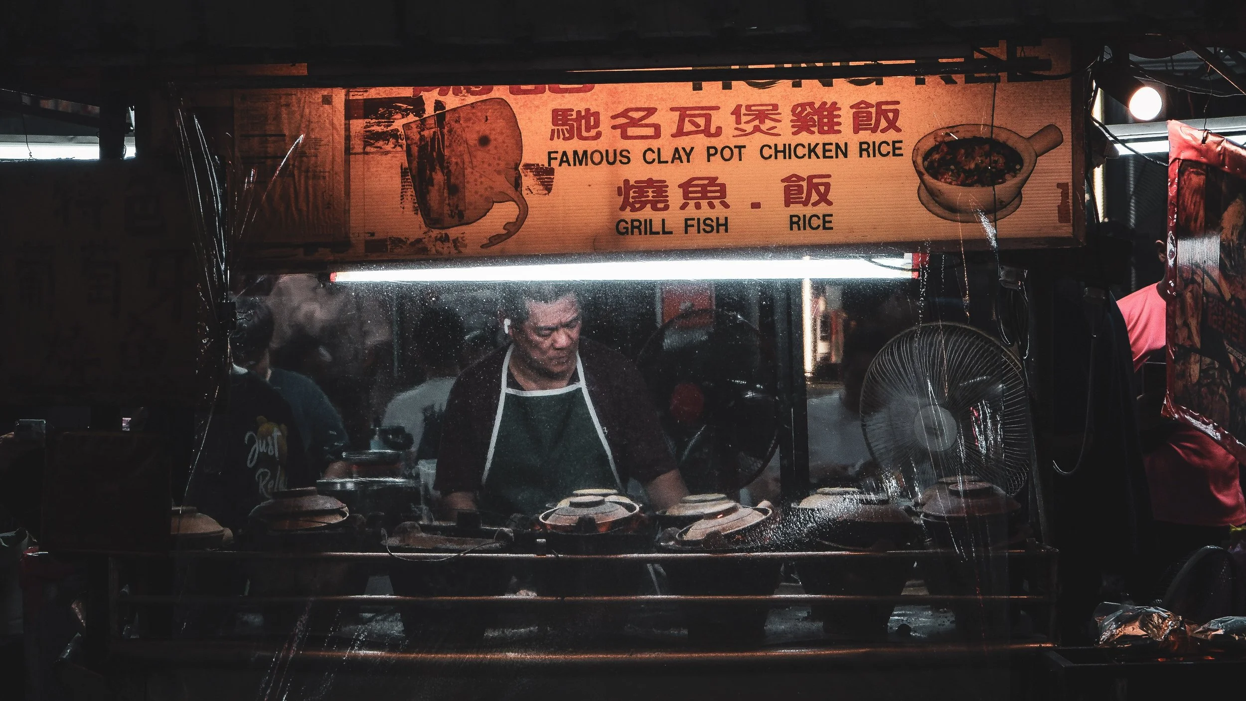 Nighttime street food stall with a sign in Chinese and English advertising clay pot chicken rice, grill fish, and rice. Inside, a man wearing an apron is cooking behind a glass display with various cooking utensils, with another person nearby.