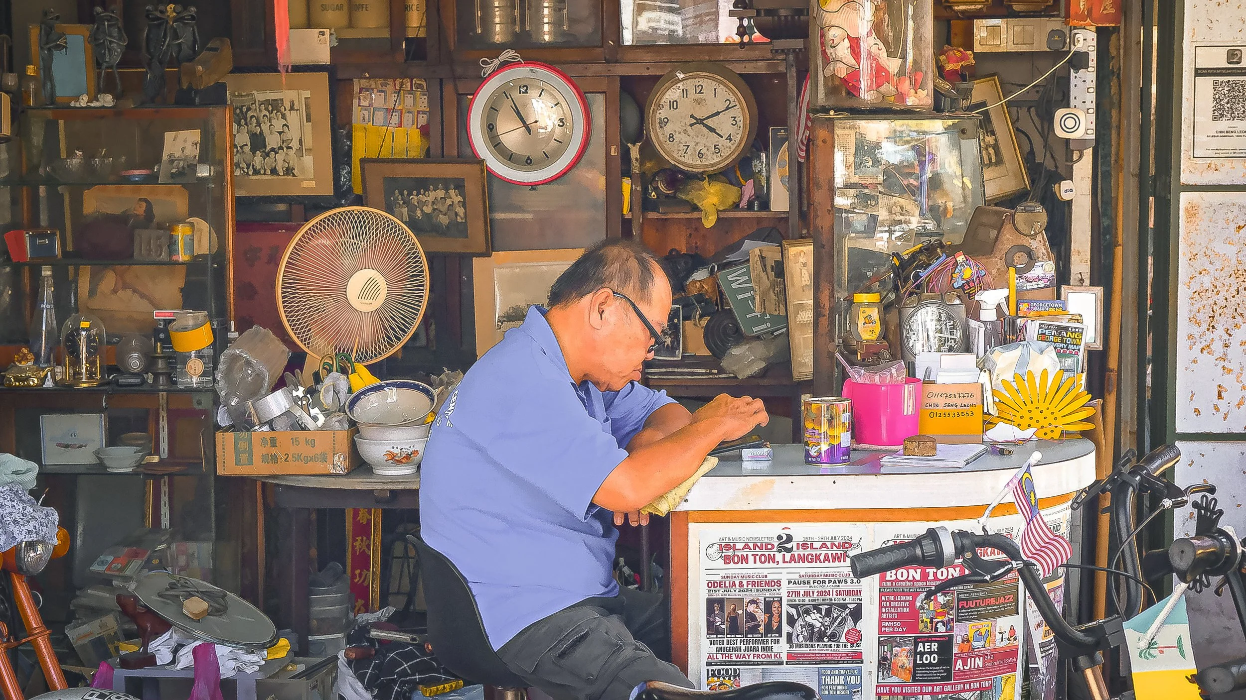A man wearing glasses and a blue shirt is sitting at a cluttered desk in a small shop or workshop, surrounded by various objects, clocks, framed photos, and souvenirs.
