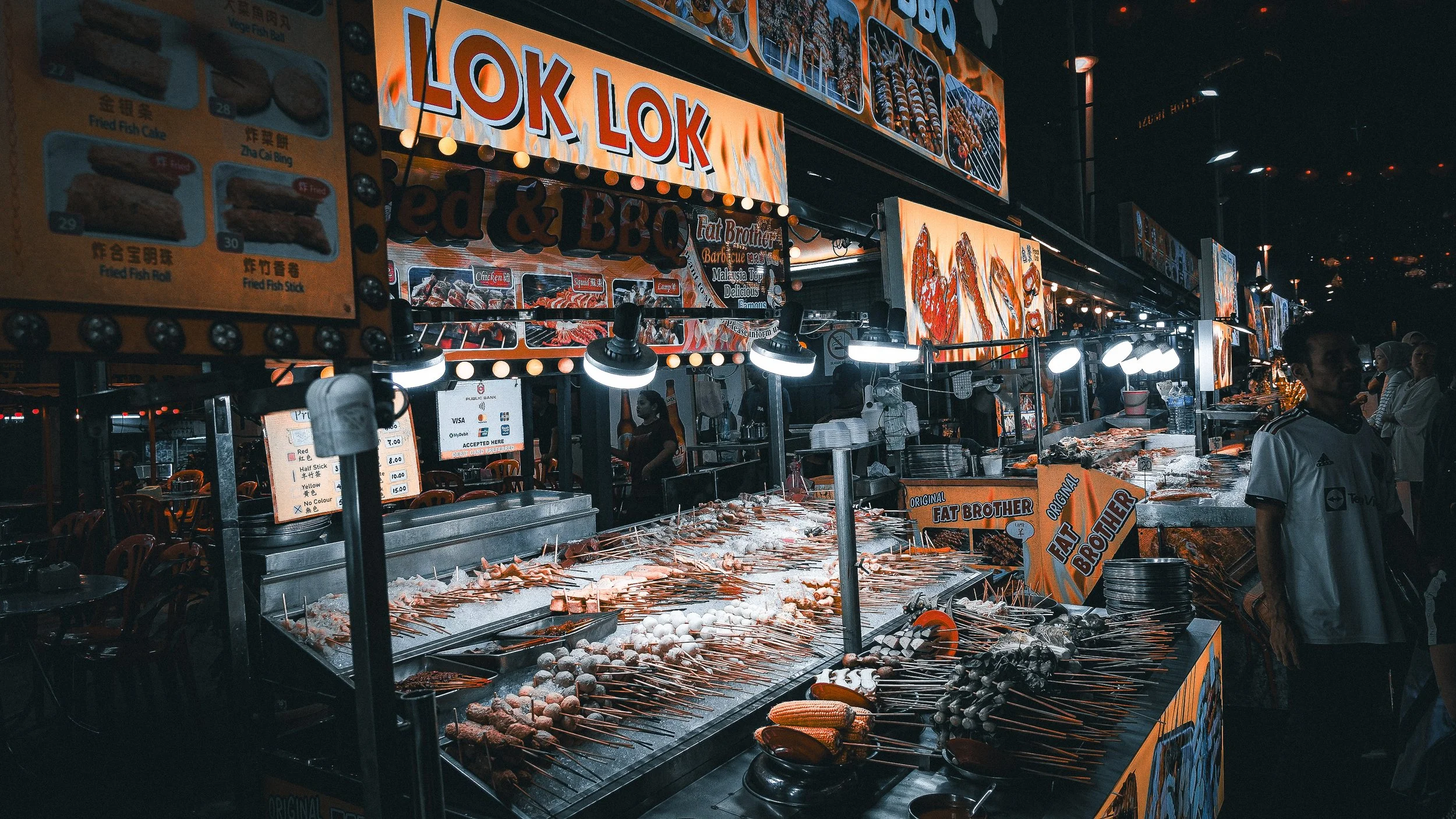 Night scene at a food stall with bright orange signage reading "LOK LOK" and parts of other signs; various skewers of raw meats, seafood, and vegetables laid out on ice, with customers and staff nearby.