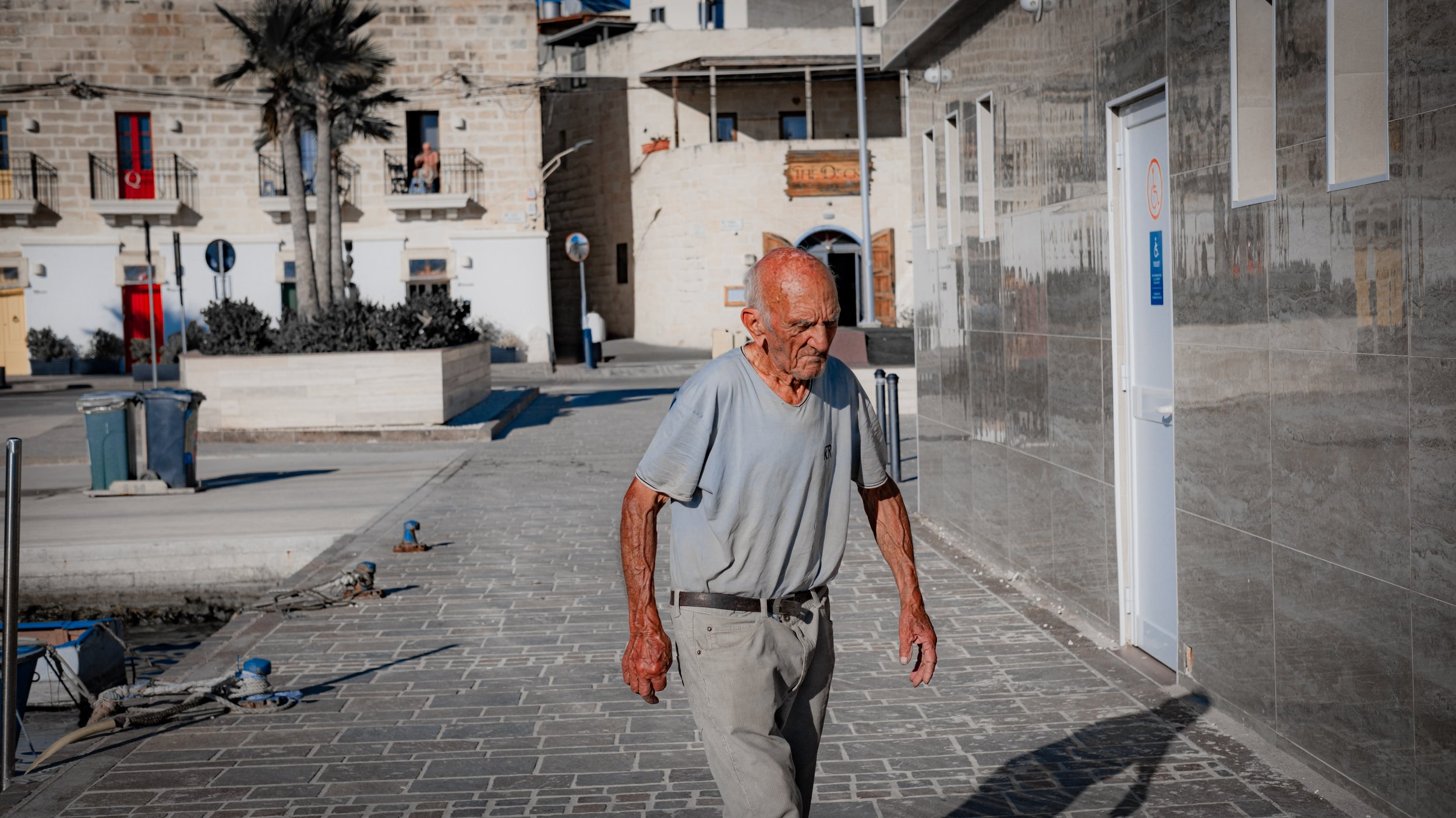 An elderly man with a shaved head walking along a paved sidewalk in an urban area with modern buildings. There is construction work and debris on the ground, and a reflective wall to the right.