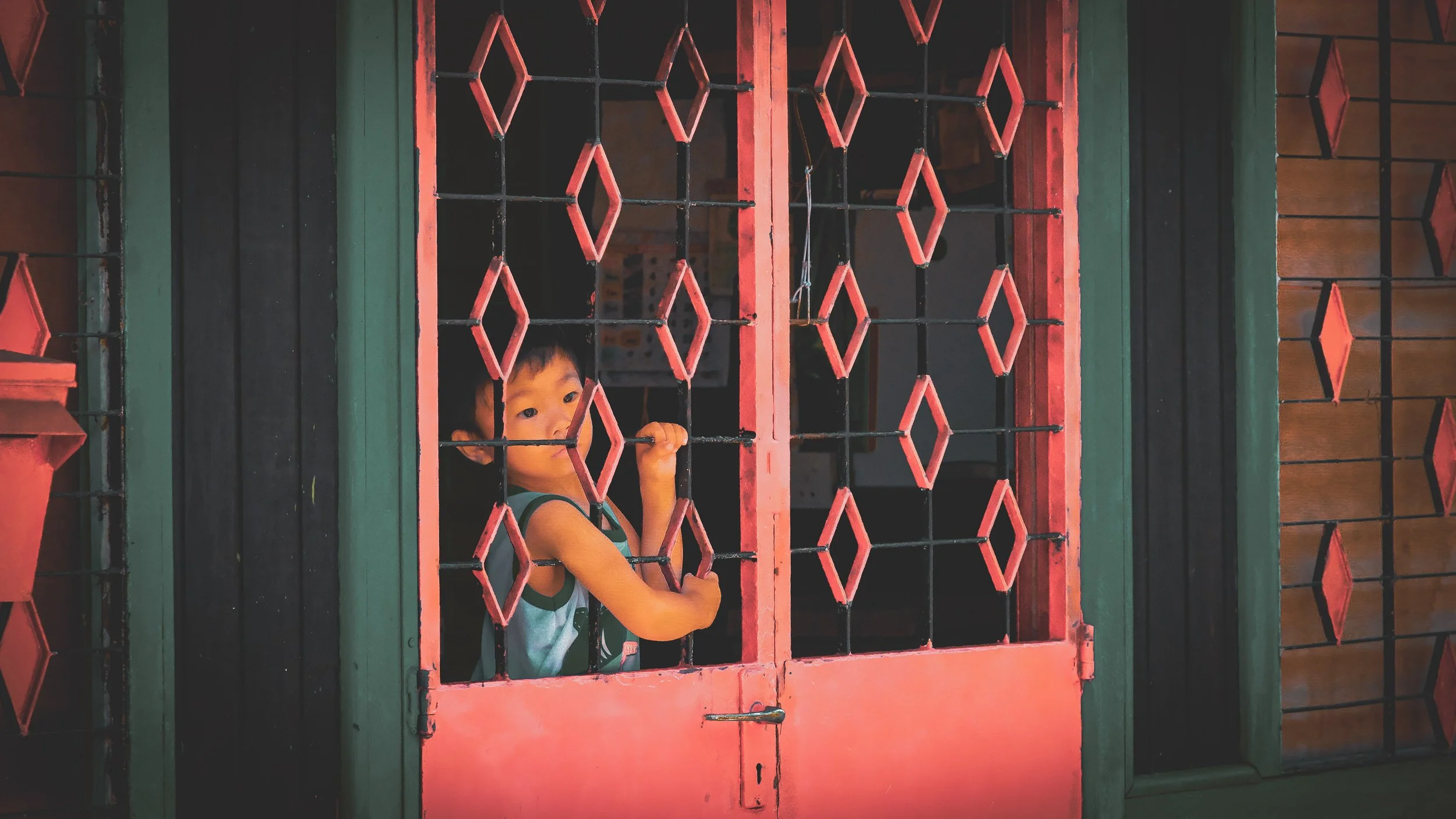 A young boy peering through red and black metal bars on a door, holding onto one of the bars with his hand, with a wooden background behind him.