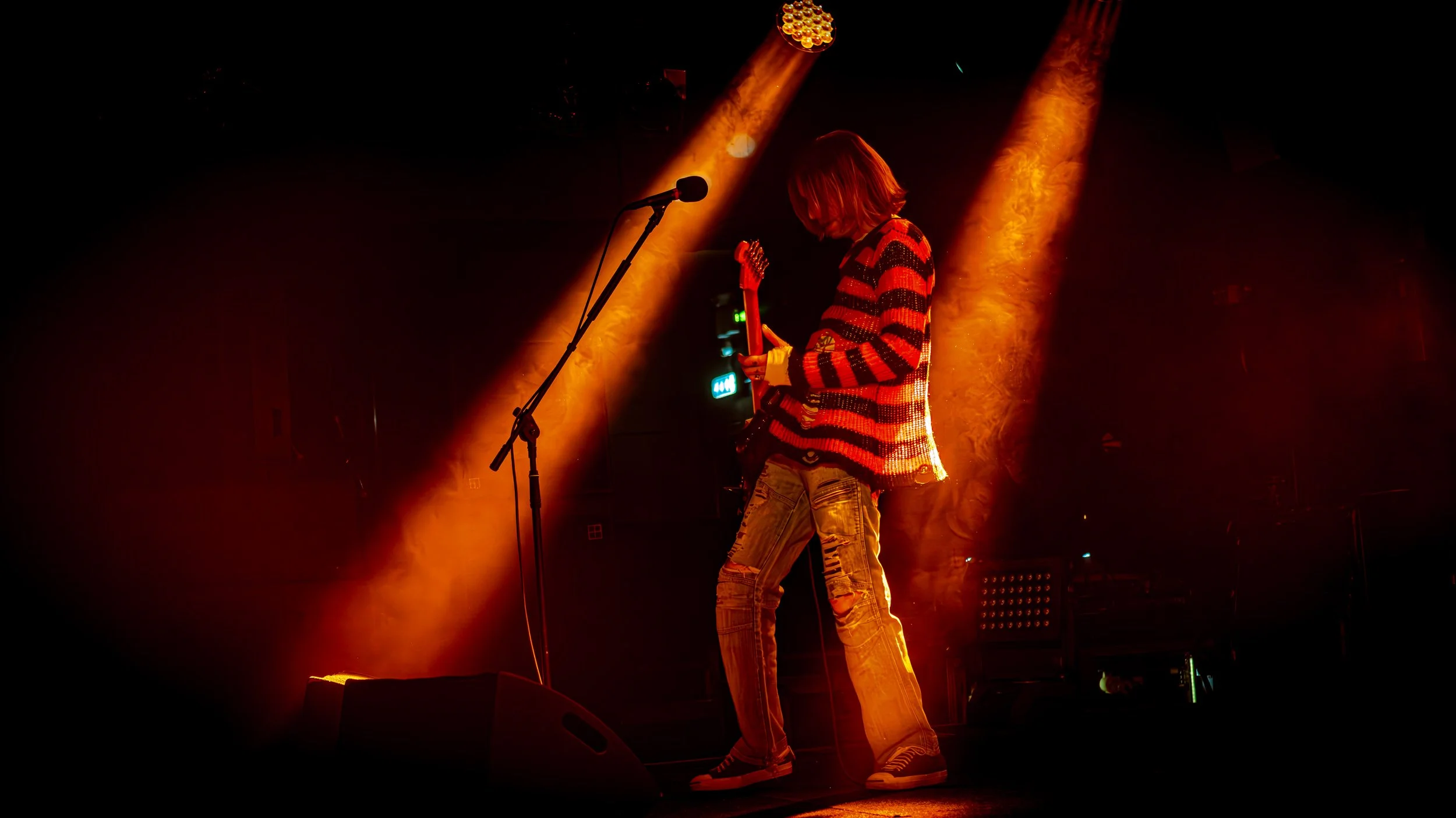 Musician performing on stage under orange and yellow stage lights, playing an electric guitar, wearing a striped sweater and ripped jeans.