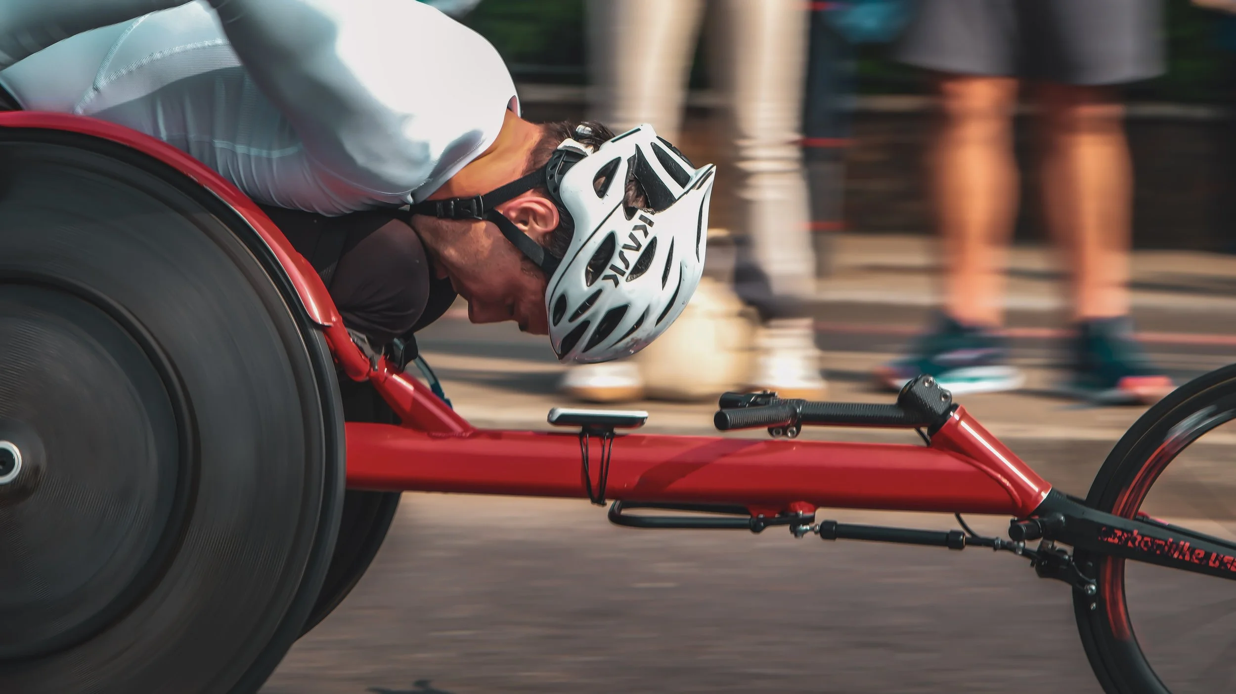A person in a racing wheelchair wearing a white helmet and white shirt, leaning forward during a race.