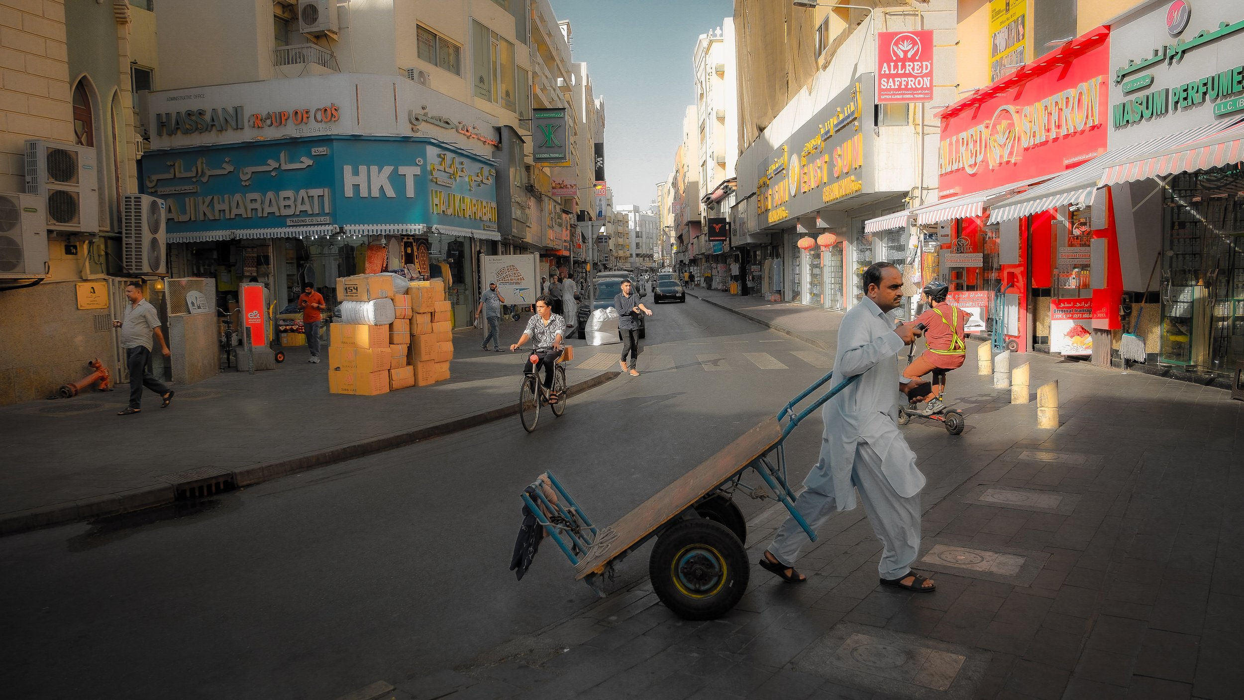 A busy city street with pedestrians, cyclists, and a worker transporting goods with a hand truck. Shops with signs, including one for saffron, line the street under multi-story buildings.