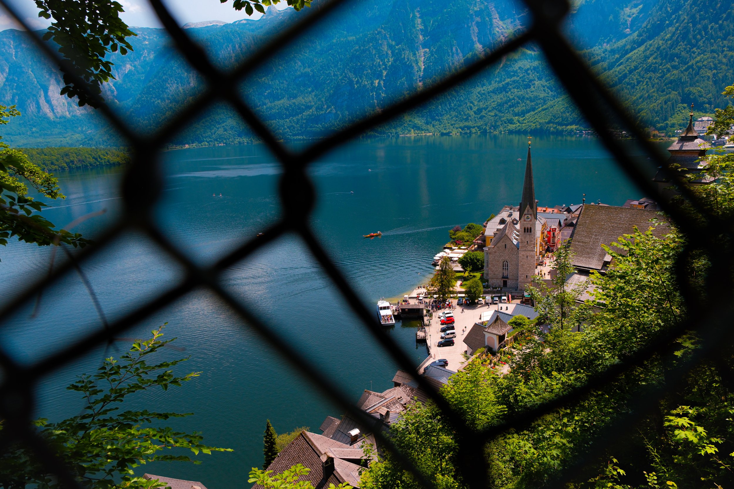 View of a large lake surrounded by green mountains, with a small town on the shoreline. The photo is taken through a fence with trees in the foreground.