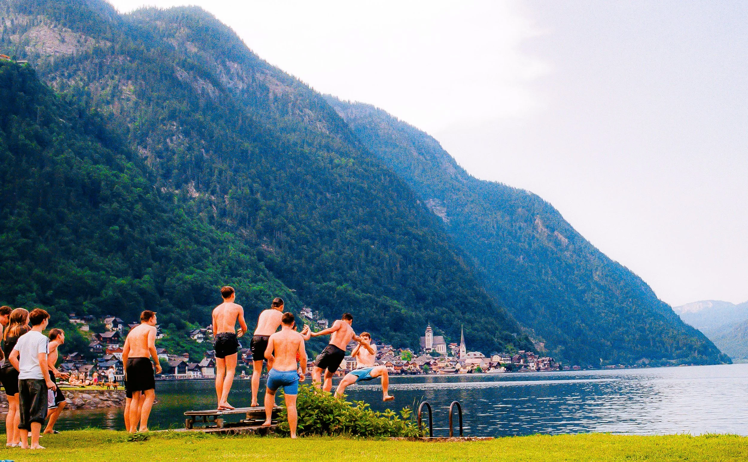 People in swimwear jumping into a lake with a mountain and a small town in the background.