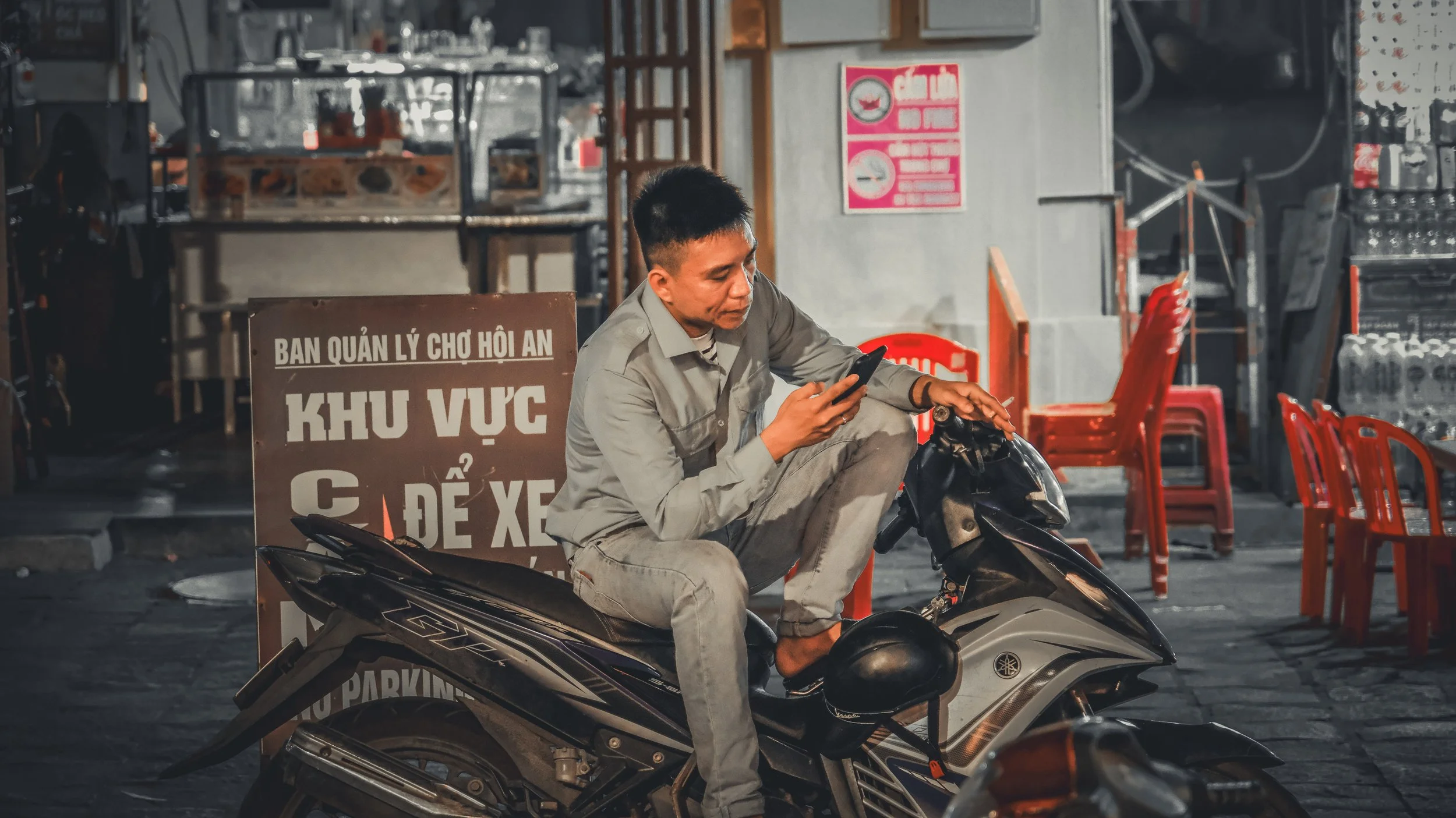 A man sitting on a Yamaha scooter in front of a sign with Vietnamese writing, next to red plastic chairs, in a dimly lit indoor setting.