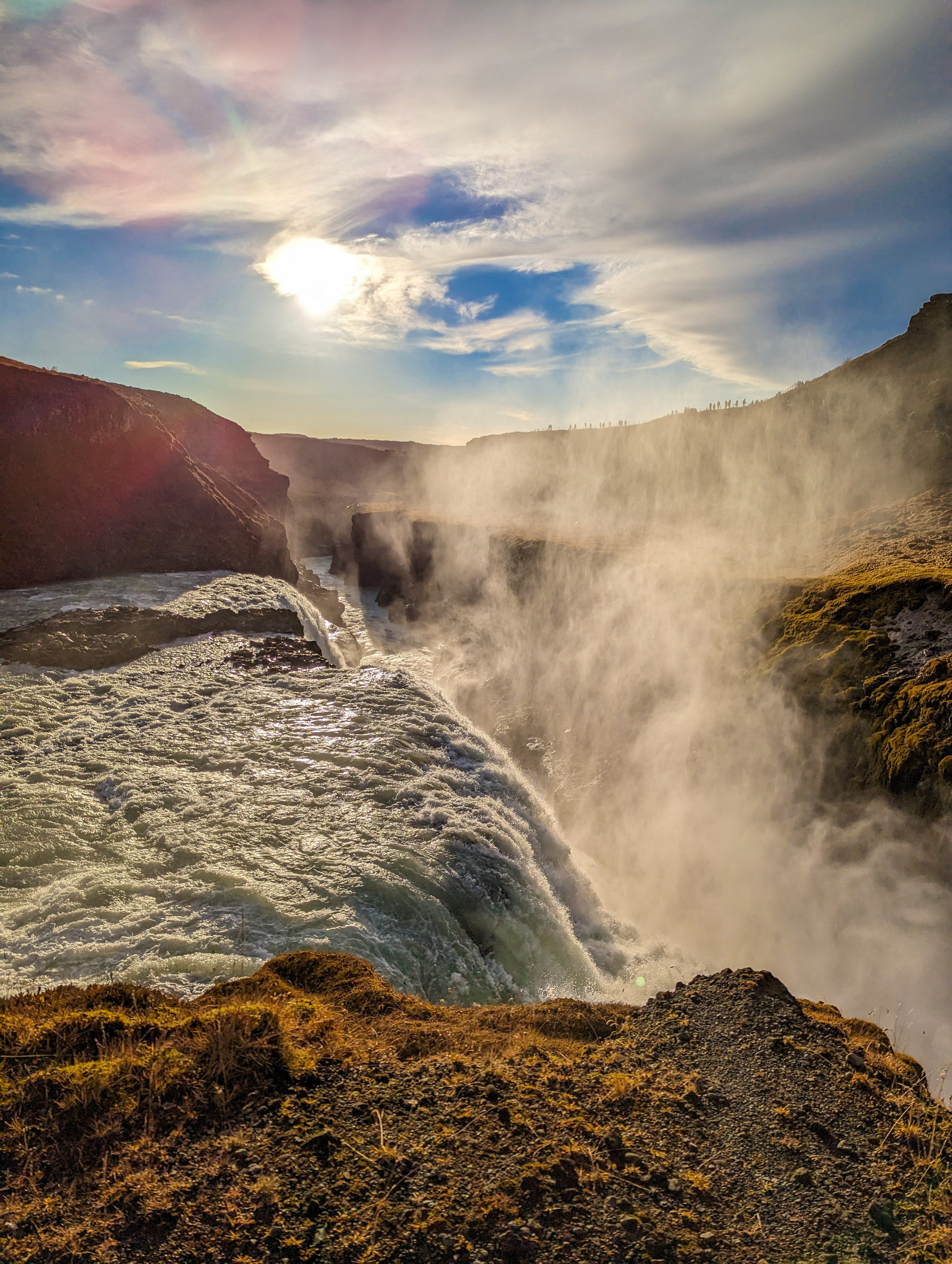 Sun shining over a canyon with waterfall and mist, rocky terrain in foreground, and a partly cloudy sky with some rainbow-like colors.