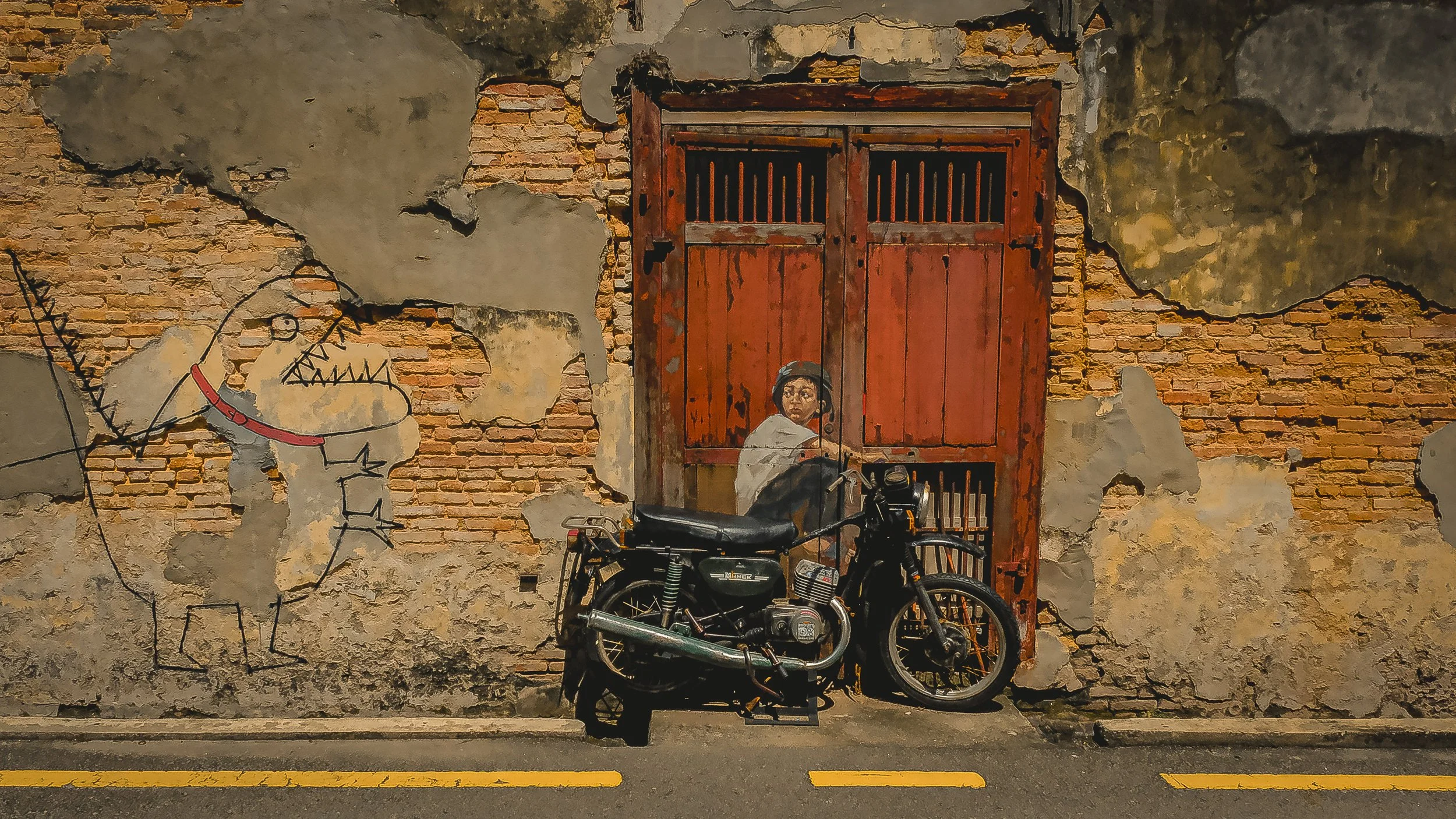 A weathered brick wall with peeling paint and mural art depicting a woman with dark hair, wearing a helmet, sitting beside a red gate. A black motorcycle is parked in front of the wall.