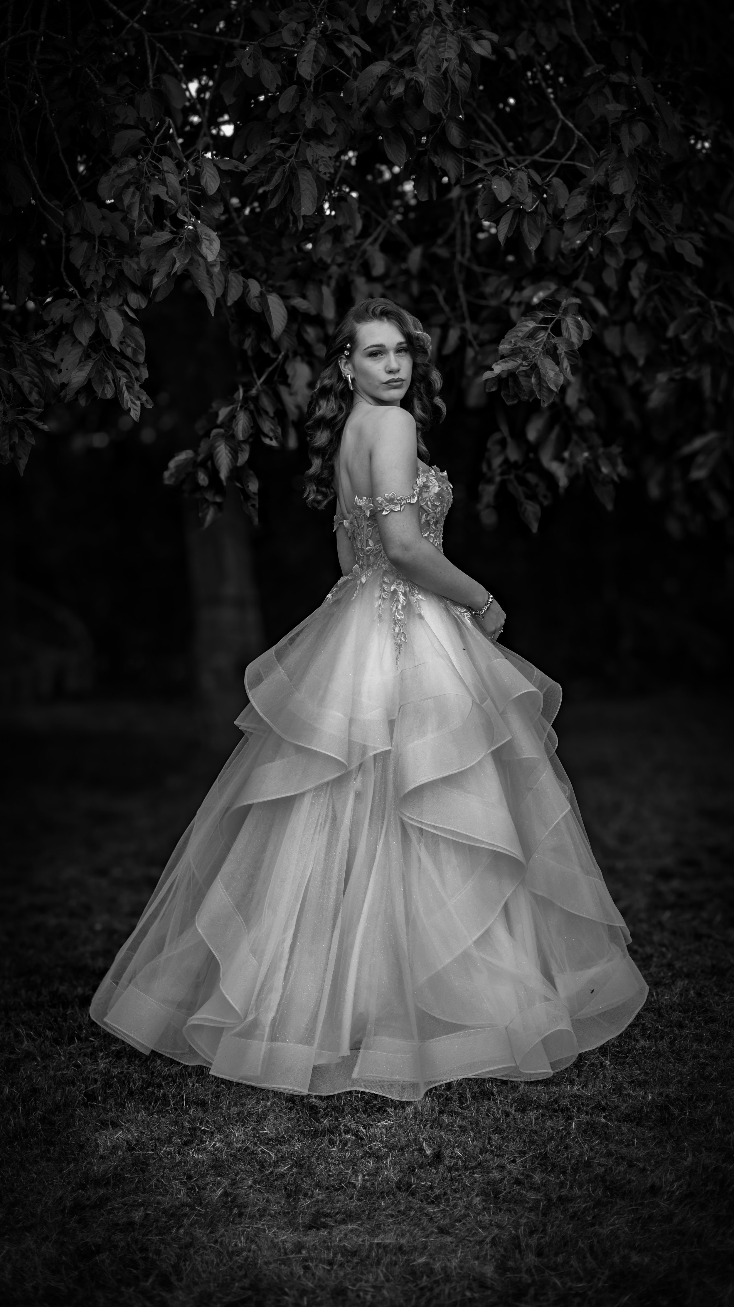 A woman in a wedding dress standing outdoors beneath a large leafy tree, black and white photograph.