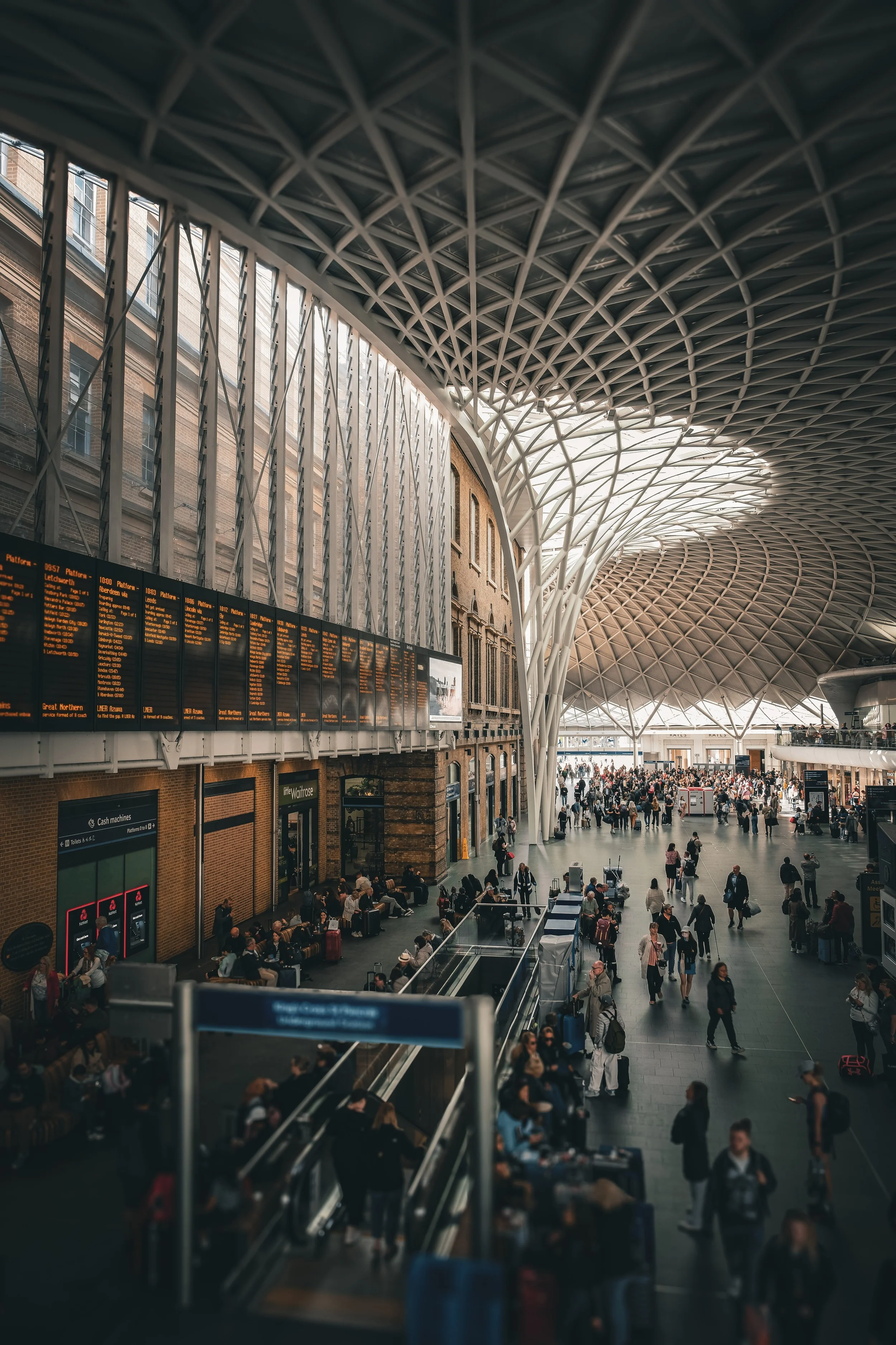 Interior of a busy train station with a modern, curved ceiling and large glass windows, filled with travelers, seating areas, and digital departure boards.