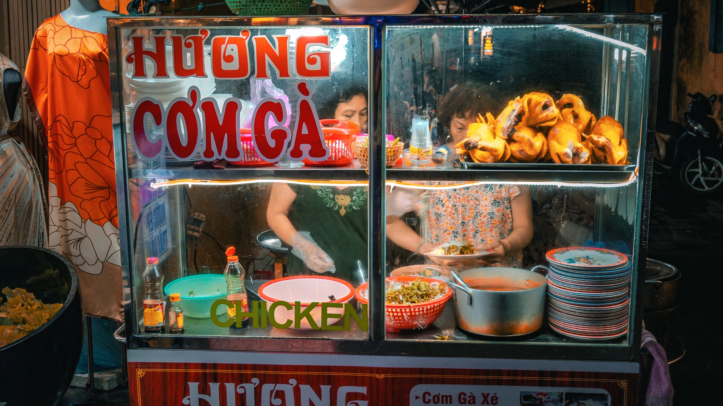 A street food stall serving chicken dishes, including a display of cooked chicken pieces, with two women preparing food inside.