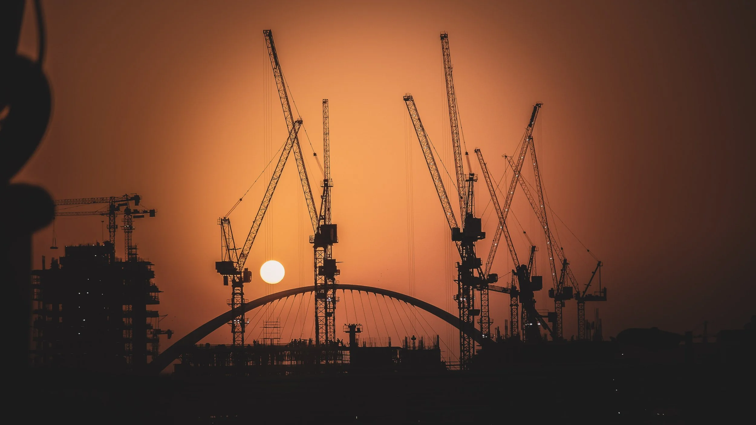 Construction cranes at sunset over a cityscape with a bridge in the background.