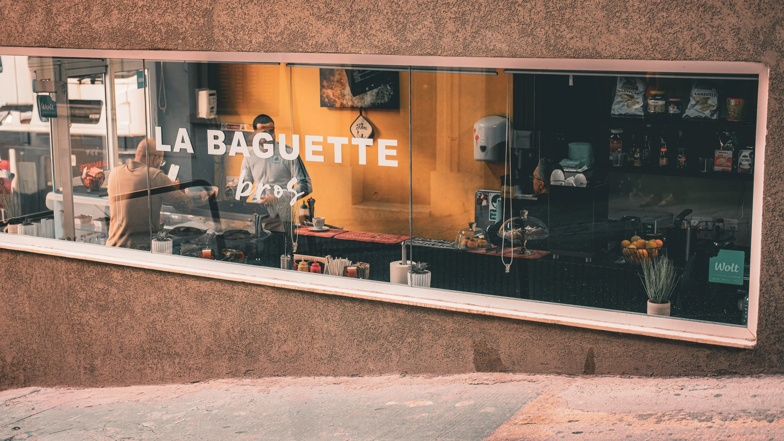 View through a restaurant window with the name 'La Baguette' displayed, showing the interior with staff and food preparation area.