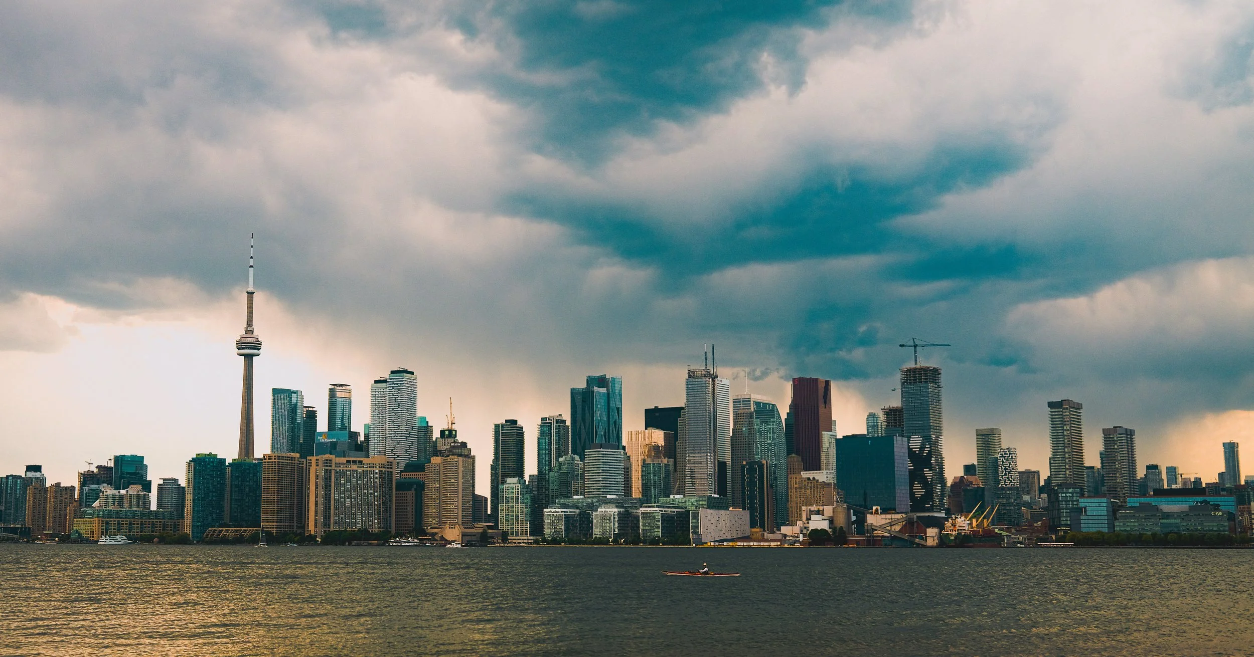 City skyline with tall buildings, including the CN Tower, under a cloudy sky, seen across a body of water with a person kayaking.