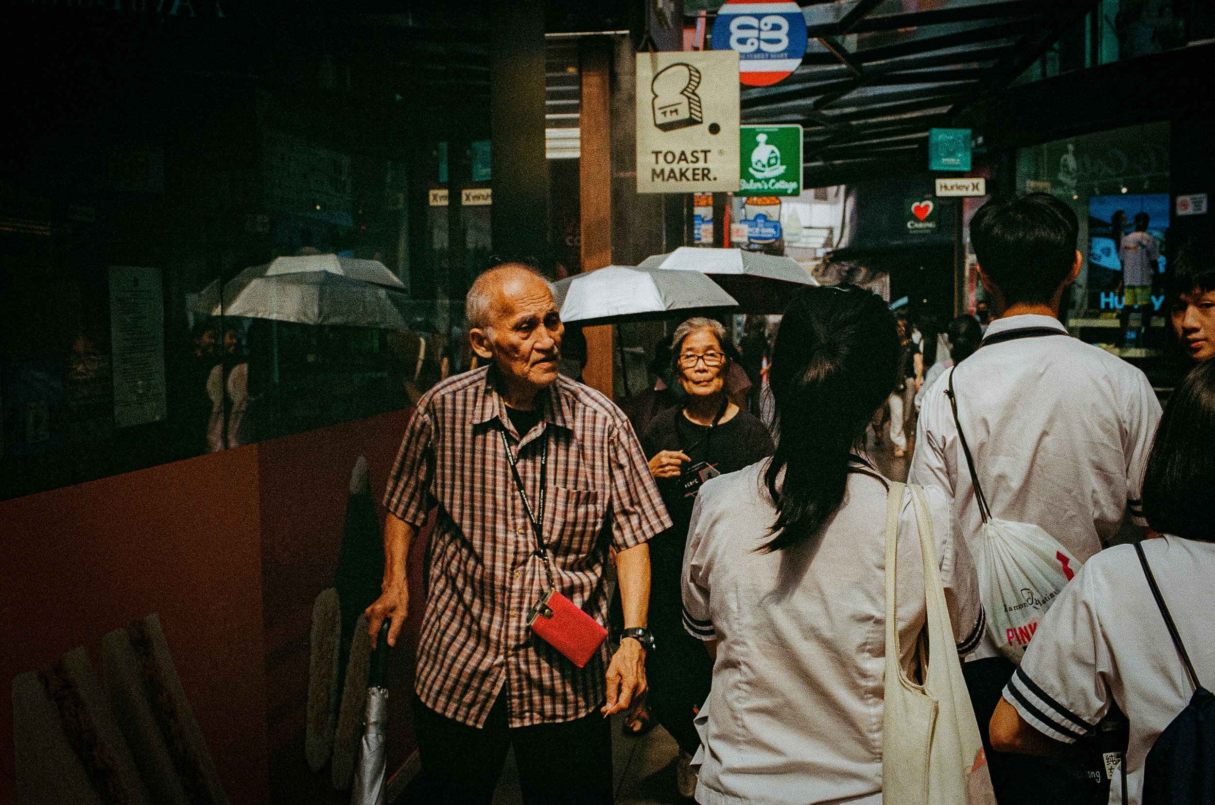 People walking on a busy street during daytime, holding umbrellas, with various signs and storefronts visible in the background.