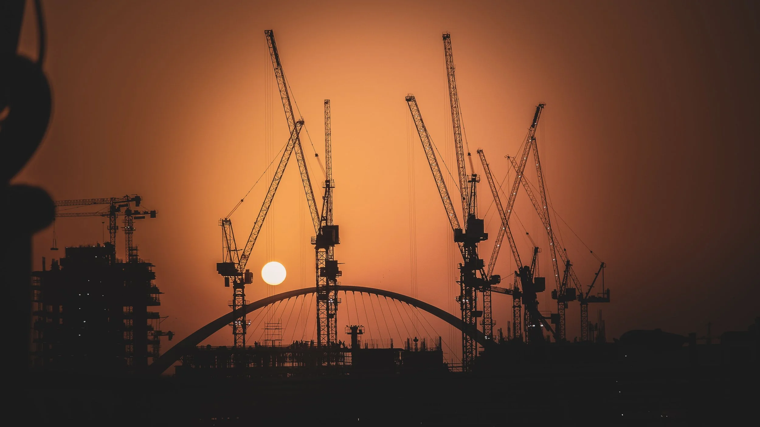 Construction cranes at sunset over a cityscape with a bridge in the background.